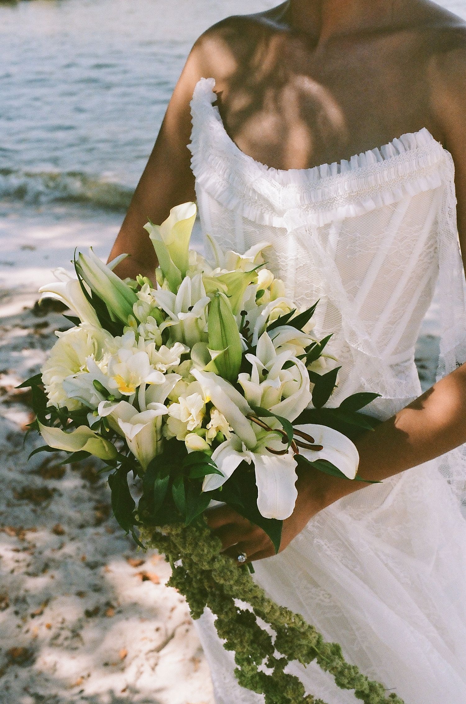 Bride in white strapless gown holding elegant bouquet of white lilies on a sandy beach.