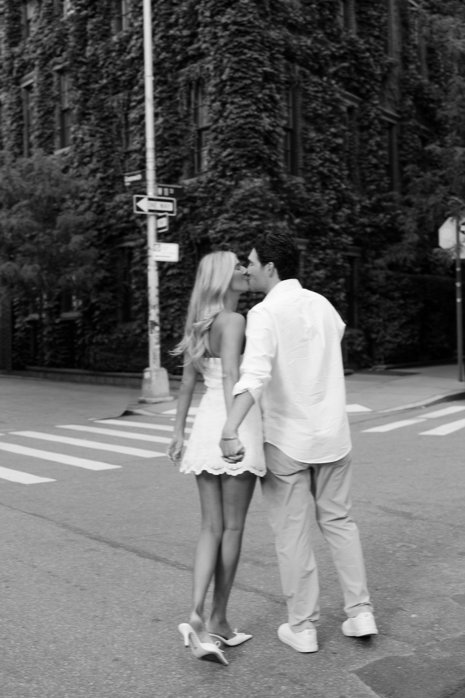 Black and white photo of a couple kissing while holding hands on a crosswalk, woman in white dress and heels.