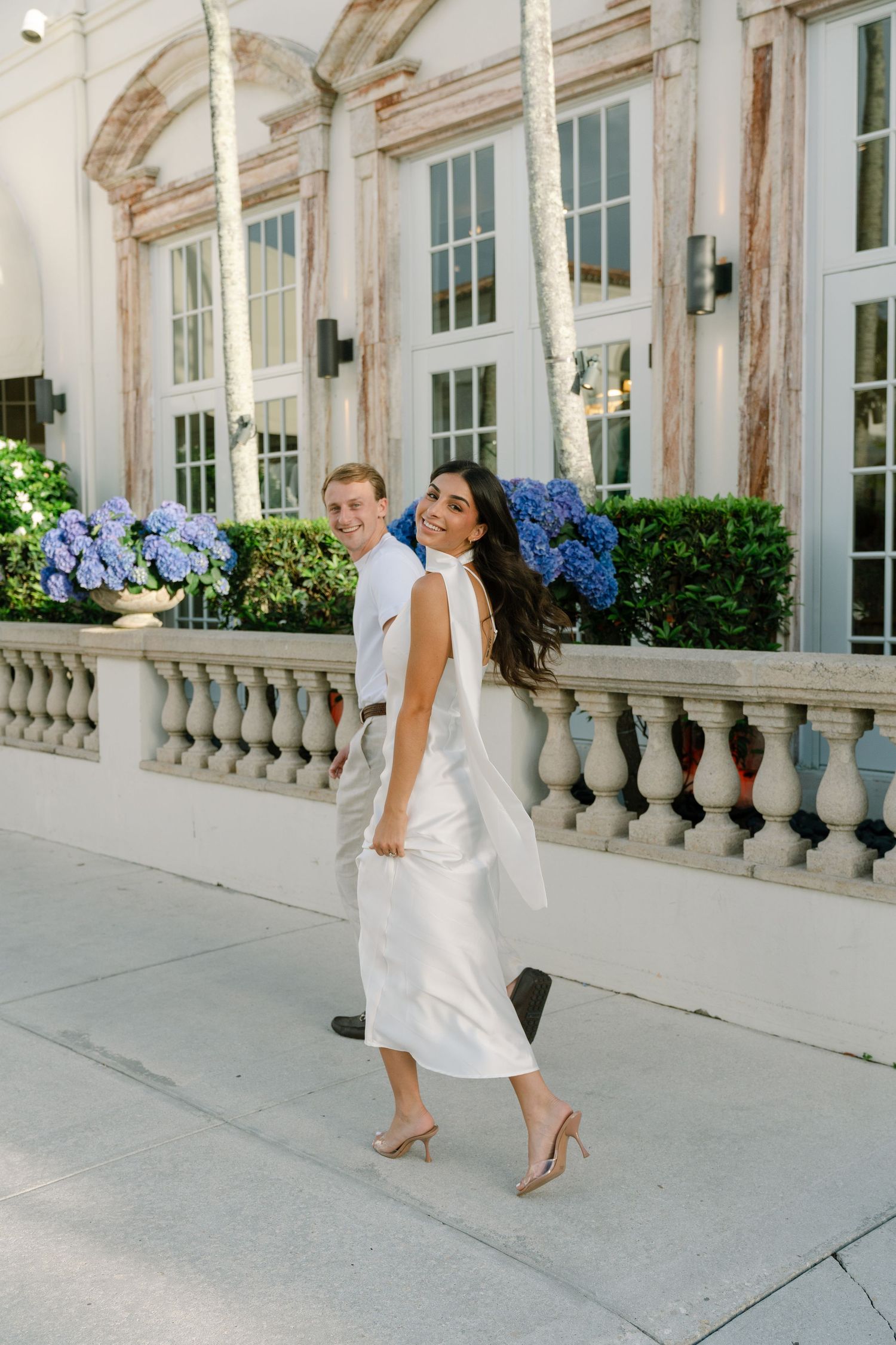 A smiling bride in a white satin midi dress walks with her groom near a elegant building with blue hydrangeas.