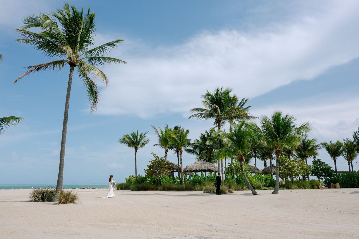 Bride in white gown walking on sandy beach with tall palm trees and gazebo under blue sky.