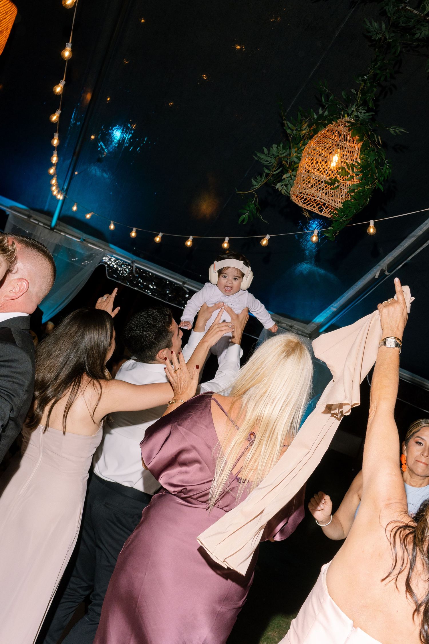 Wedding guests lifting baby on dance floor under string lights at lively indoor reception.