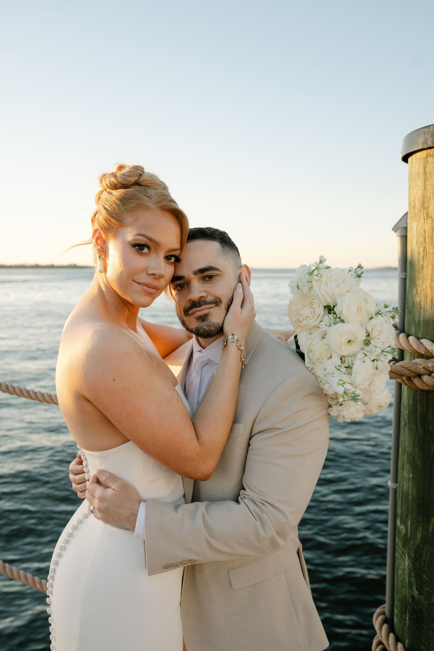 Romantic wedding couple embraces on a wooden dock by the ocean at sunset with white floral decorations.