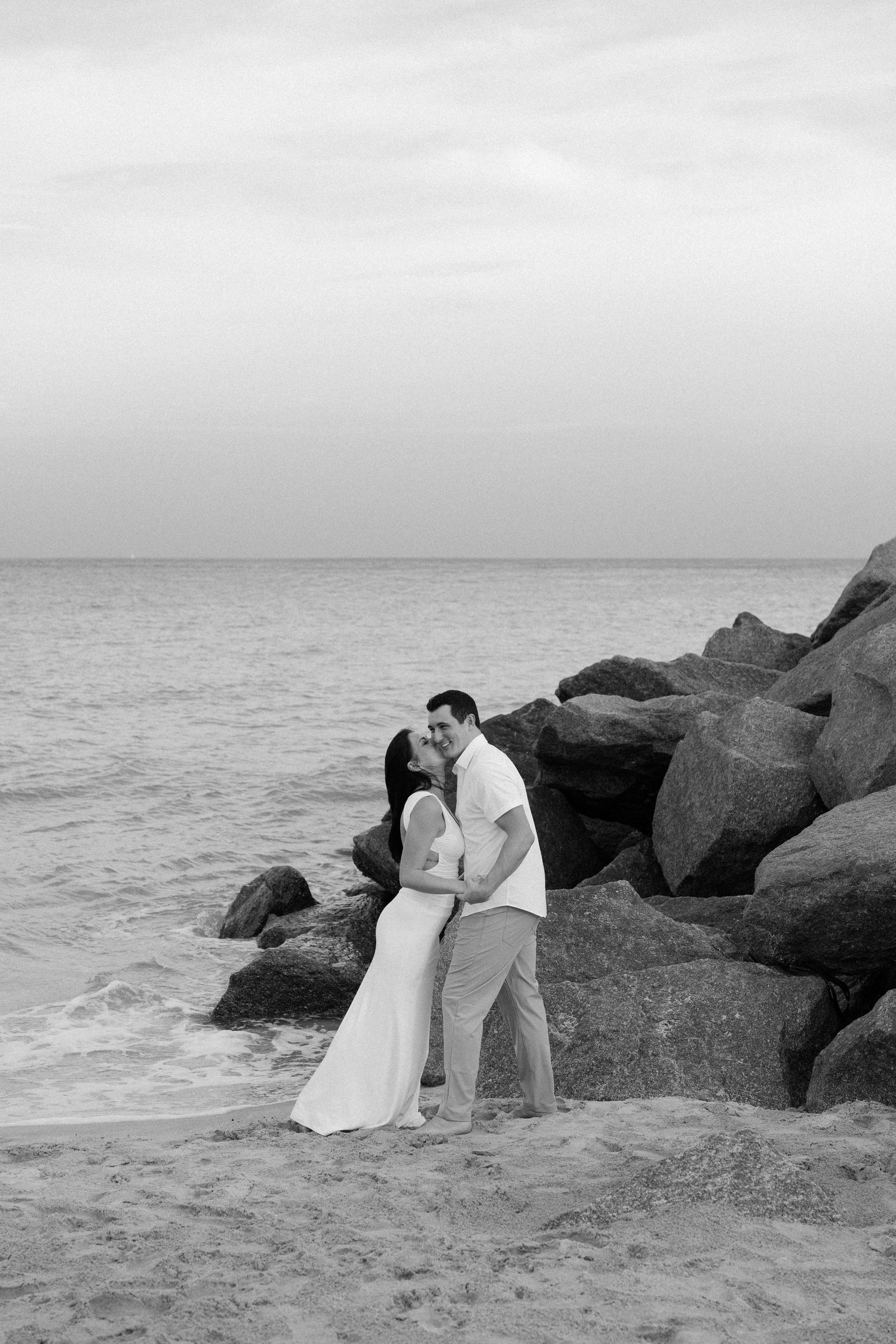 A romantic couple poses on rocky coastal outcrop in black and white photo series.