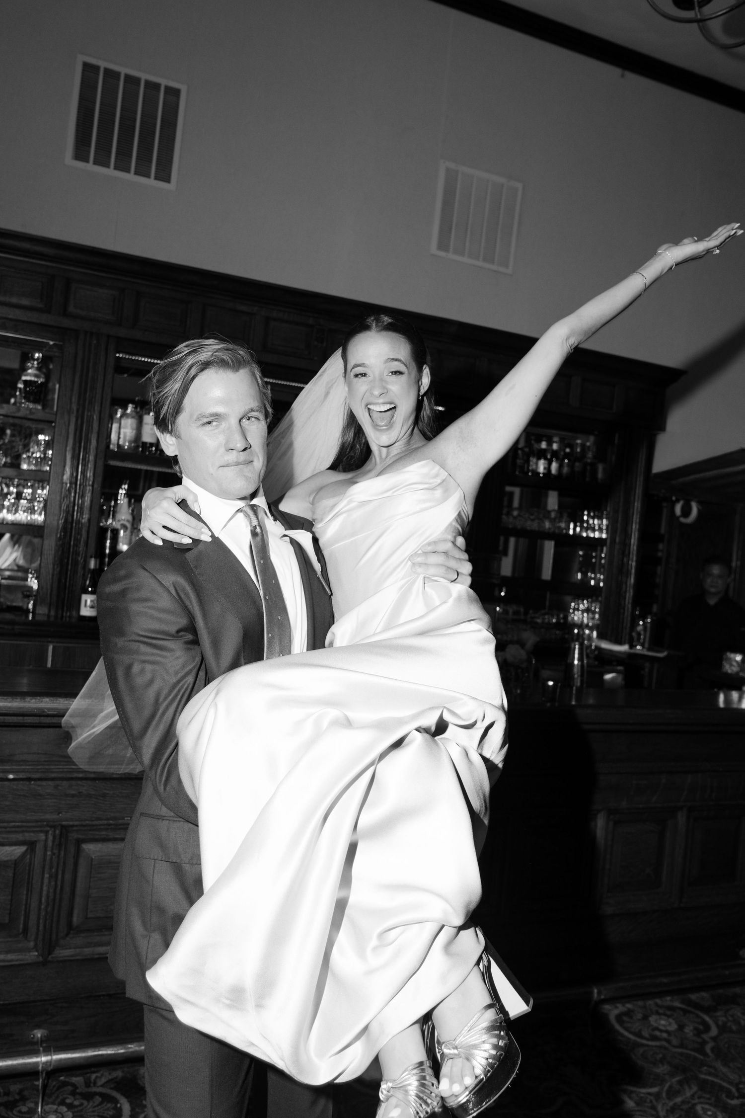 Groom carries joyful bride in white gown and veil at elegant wedding reception venue in black and white photo.