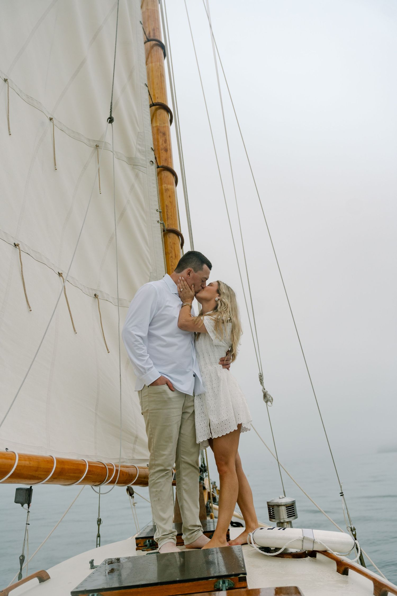 A couple shares a romantic kiss on the deck of a classic sailboat on calm waters.