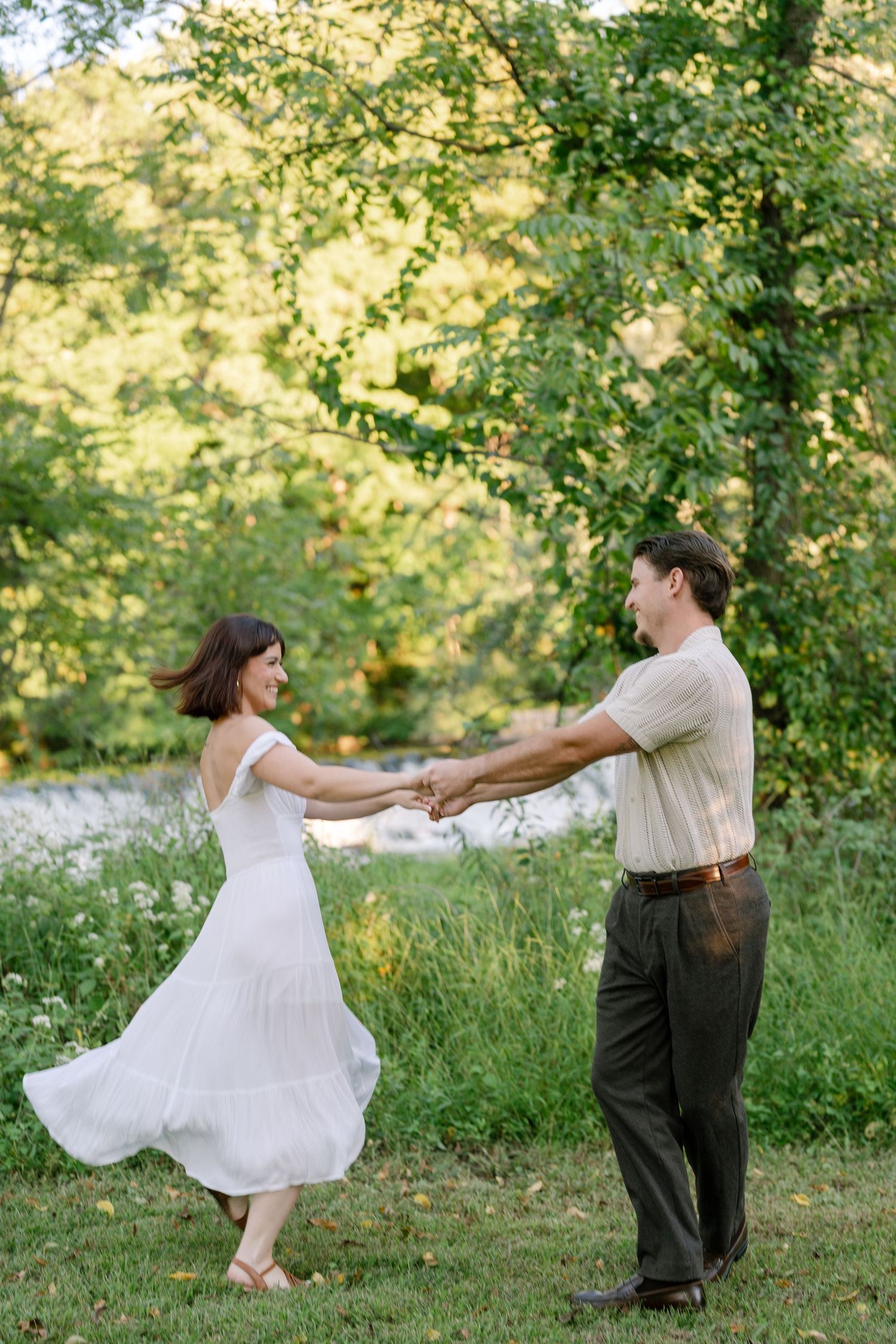 Couple dancing outdoors in lush green park, woman in white dress twirling, man in striped shirt and olive pants.