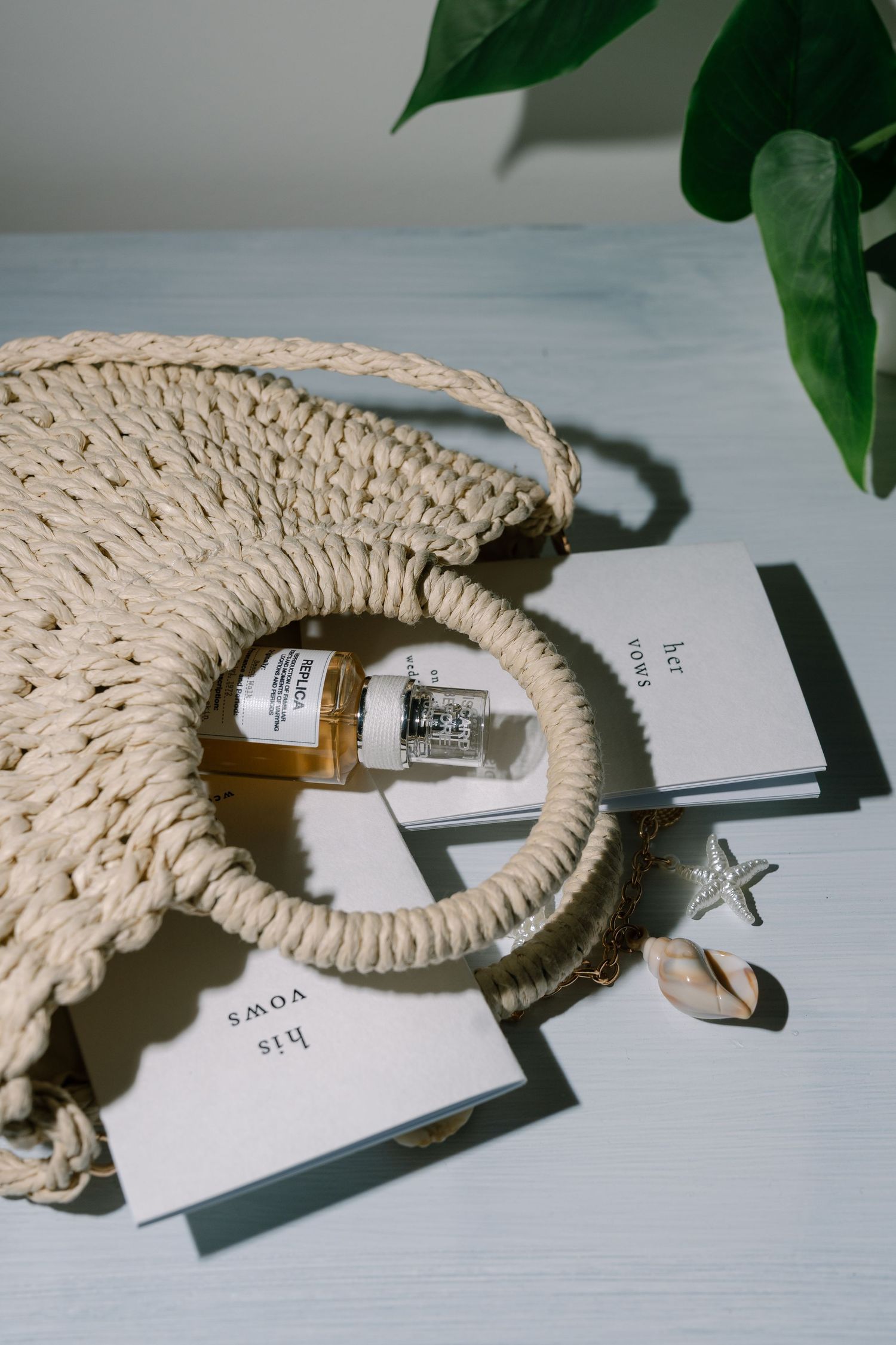 Woven straw circle bag with perfume bottle, vow books, starfish, and shell on blue surface.