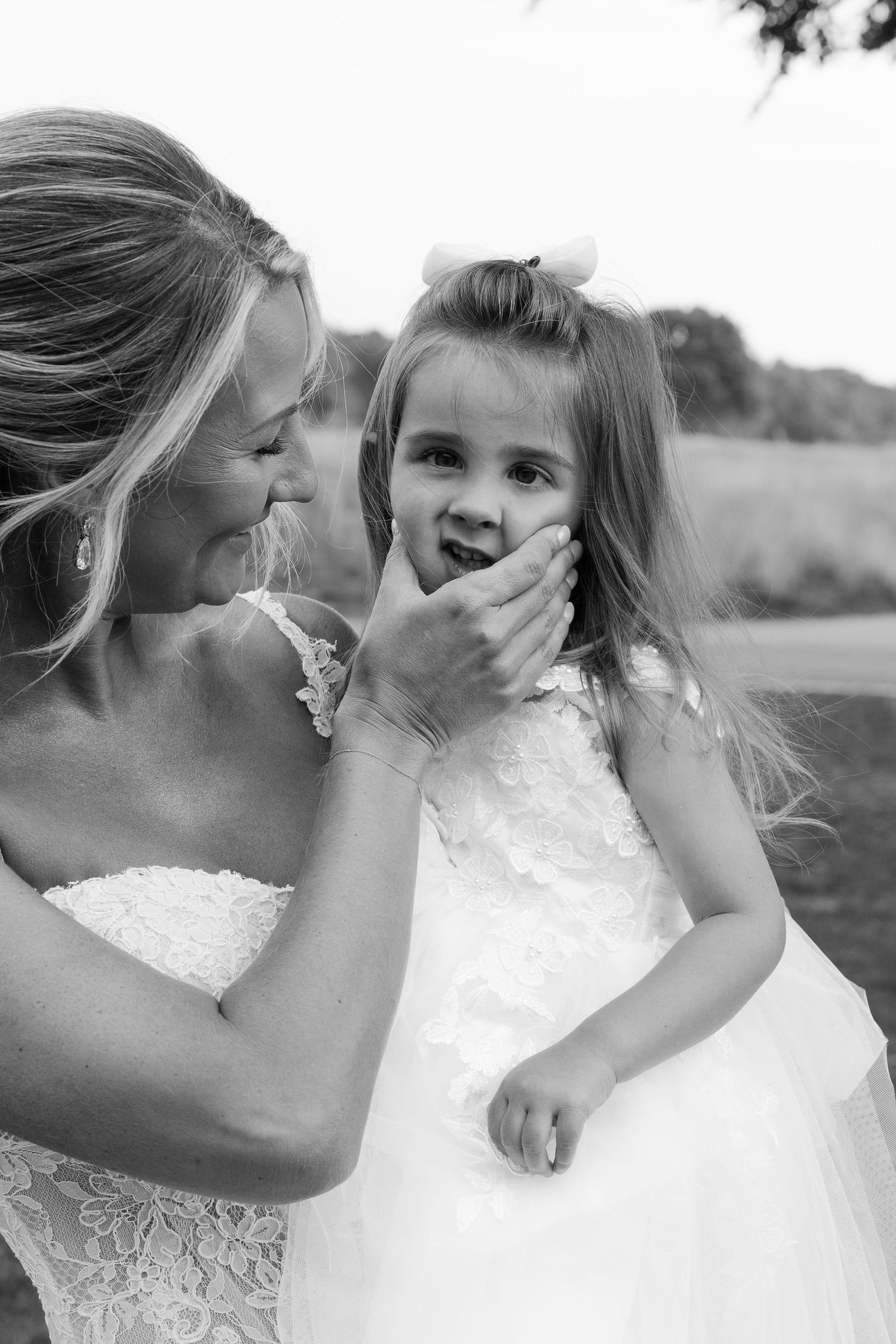 Black and white photo of a laughing bride tenderly covering a young flower girls mouth at an outdoor wedding.