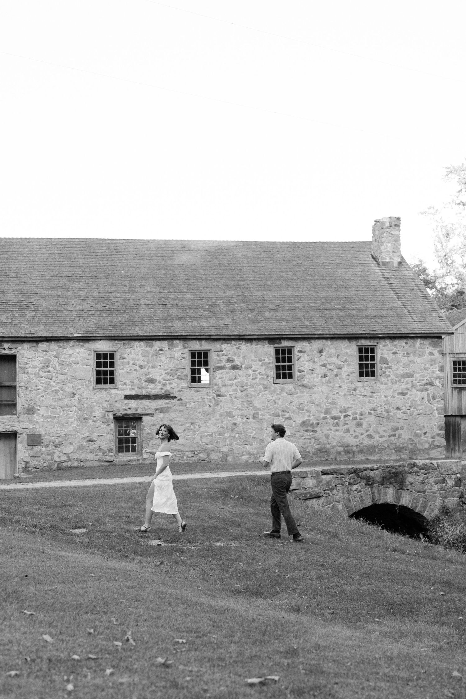Black and white photo of a couple walking near a historic stone building with arched bridge on a grassy lawn.