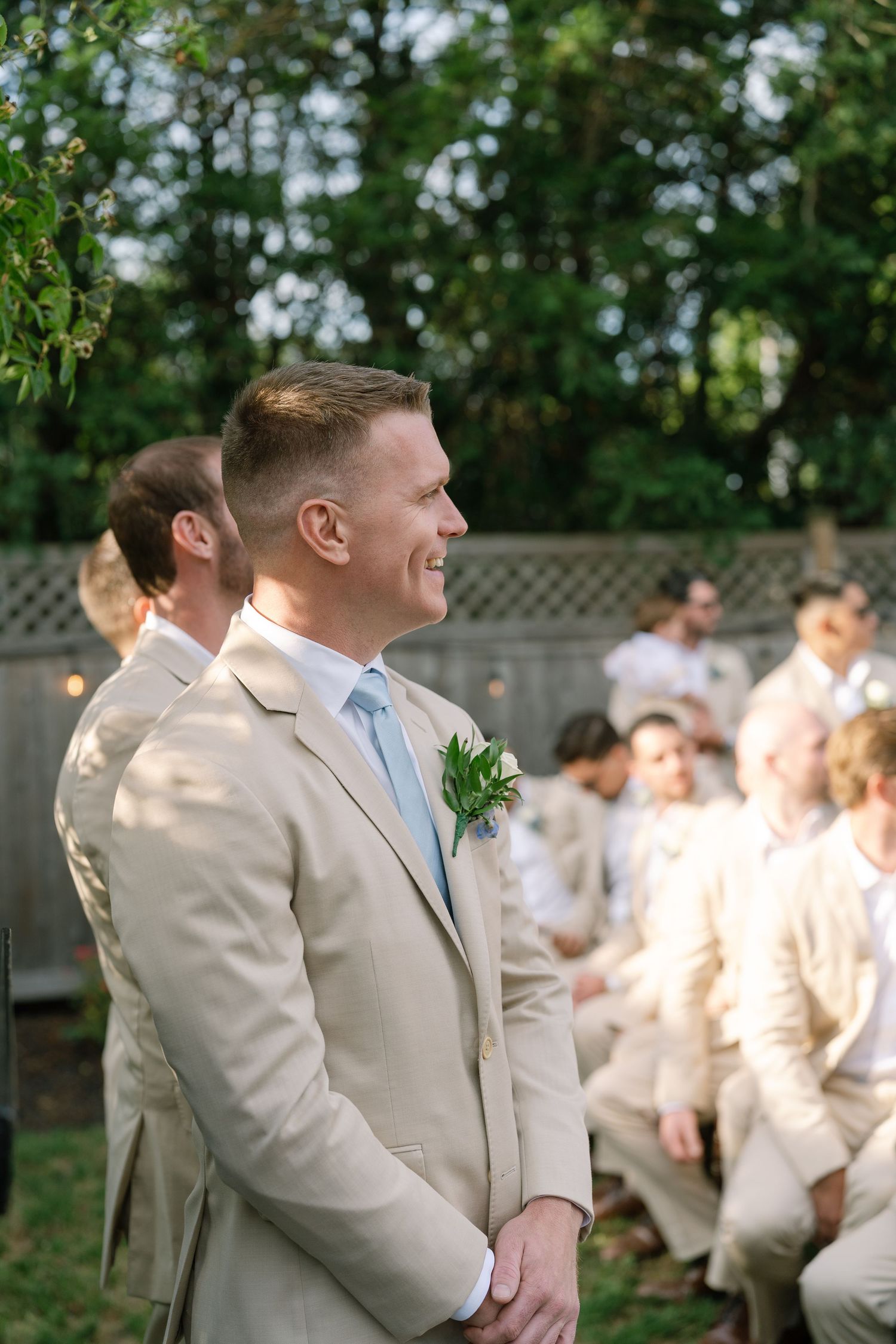 Smiling groom in beige suit with boutonniere awaiting bride at outdoor garden wedding ceremony.