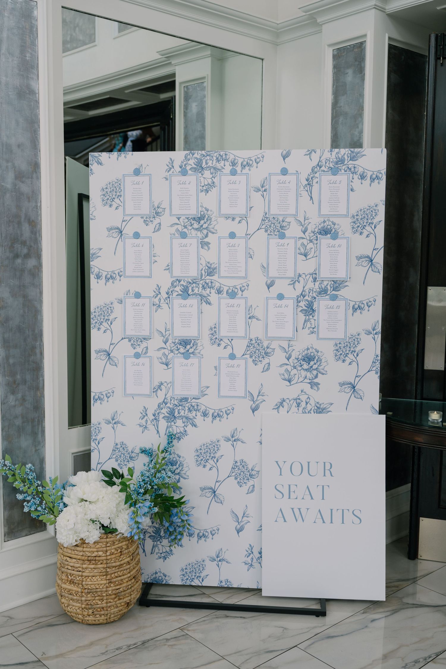 Blue floral seating chart display board with Your Seat Awaits sign and white hydrangea arrangement at wedding reception.