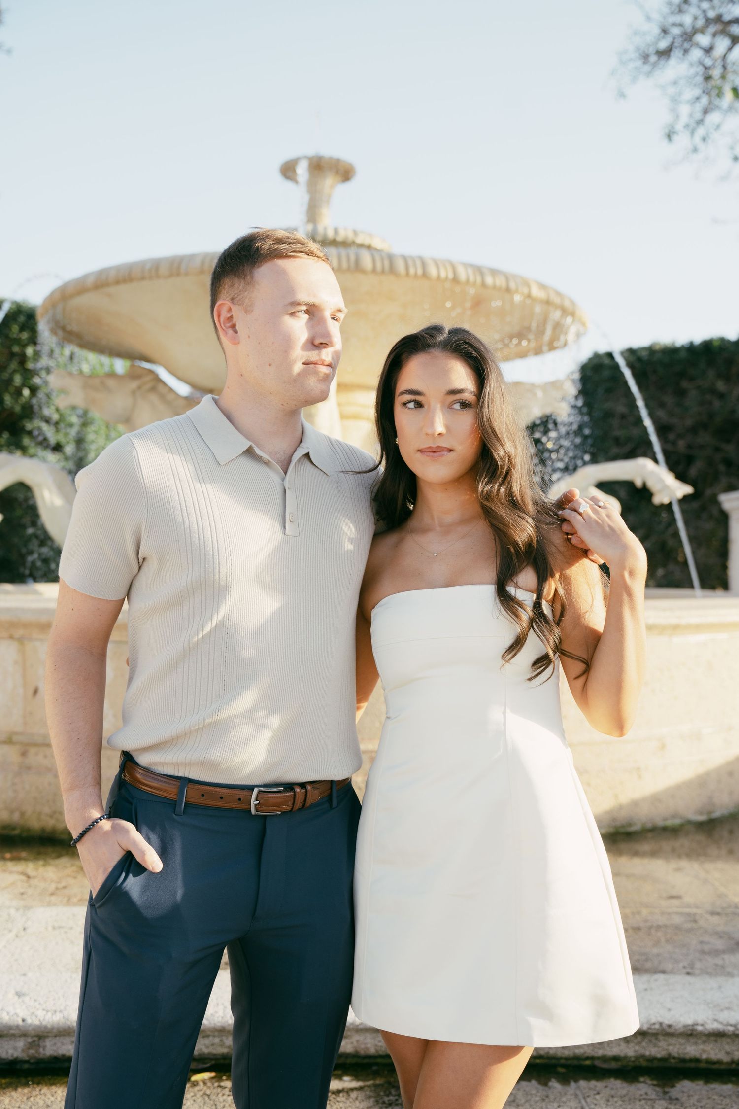 A couple in casual attire poses together outdoors during a sunlit photoshoot against a soft natural background.