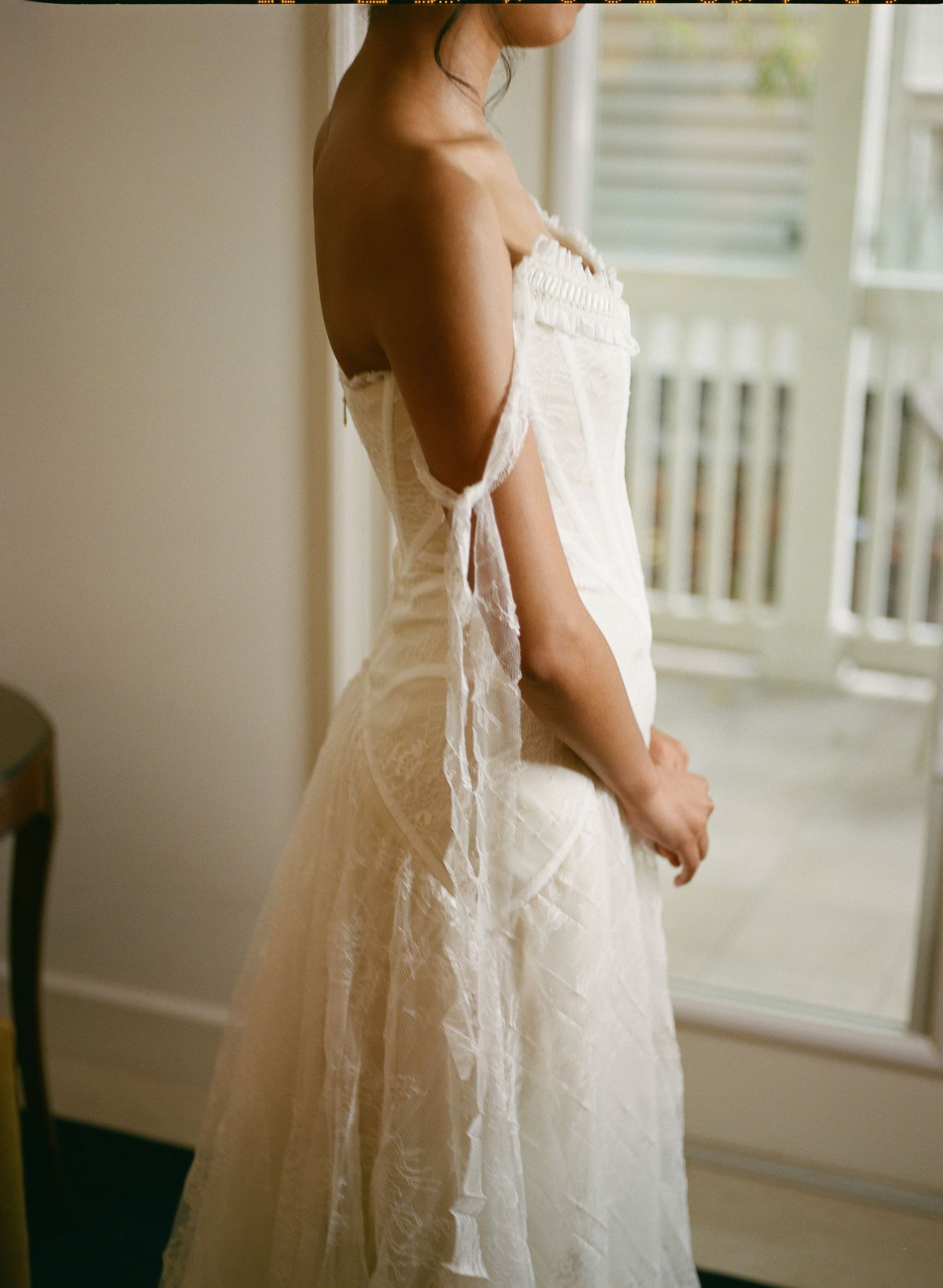 Bride in strapless lace wedding gown with open back tie detail standing by window in soft natural light.