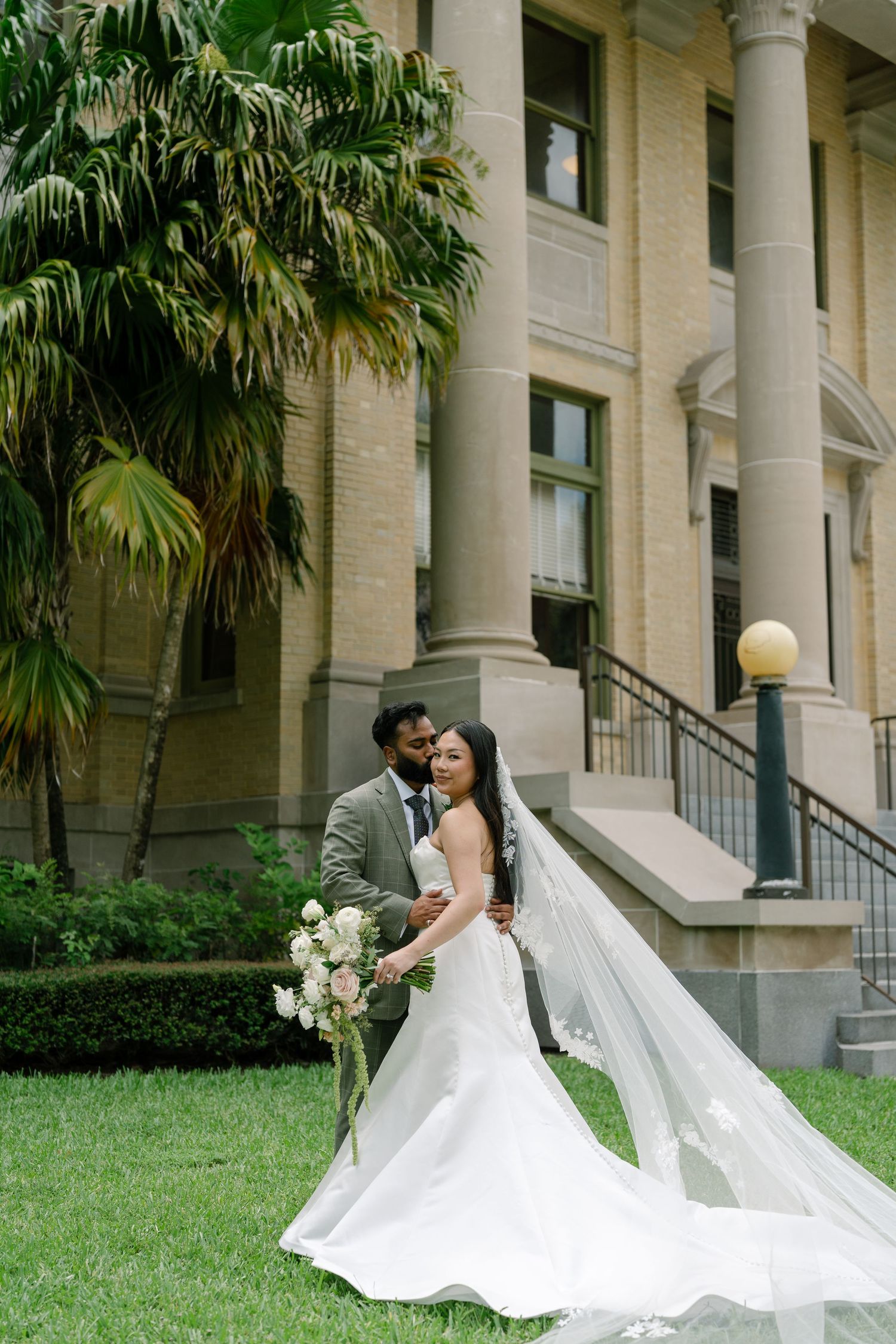 Bride and groom pose outdoors near a grand building with columns, her veil flowing gracefully as she holds a white bouquet.