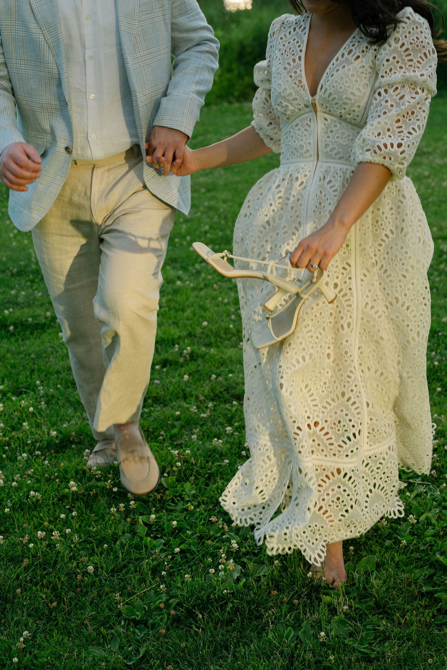 Couple holding hands running barefoot on green grass, woman in white eyelet lace dress carrying heels.
