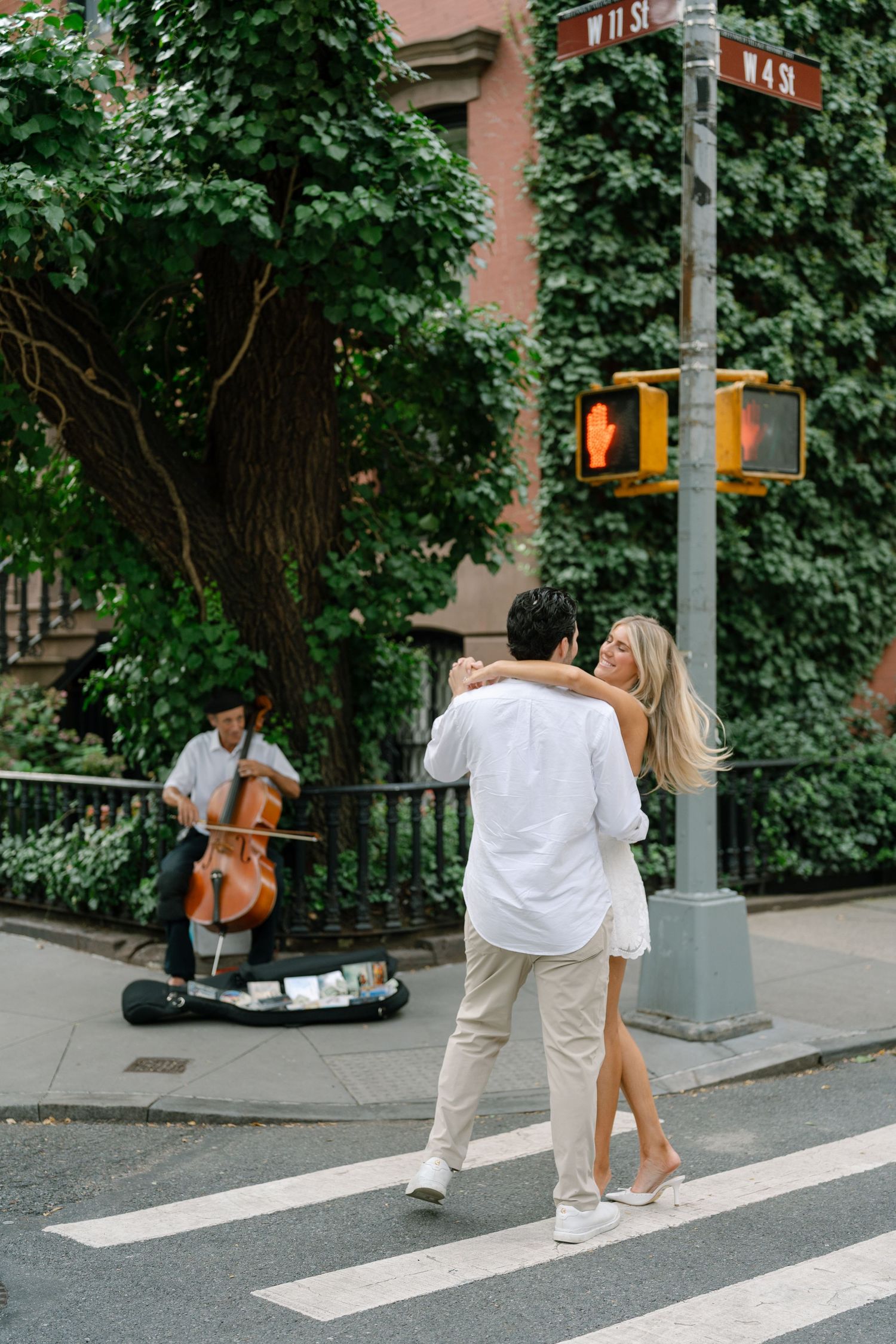 Couple dancing on NYC crosswalk at W 11th St while a cellist performs nearby amid lush green ivy.
