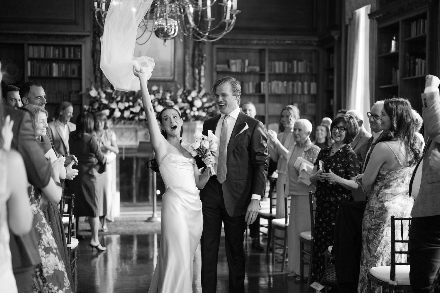 Black and white photo of joyful bride raising bouquet after wedding ceremony in elegant library venue with guests applauding.