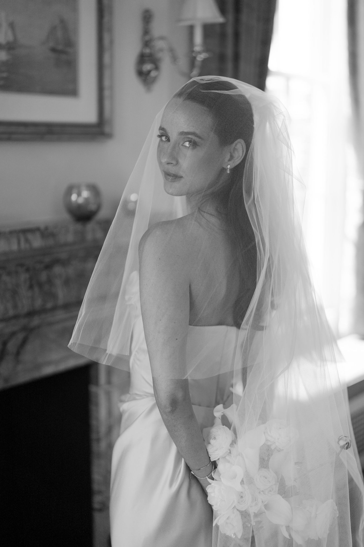 Black and white portrait of a bride in a strapless gown and veil holding a bouquet near a window.
