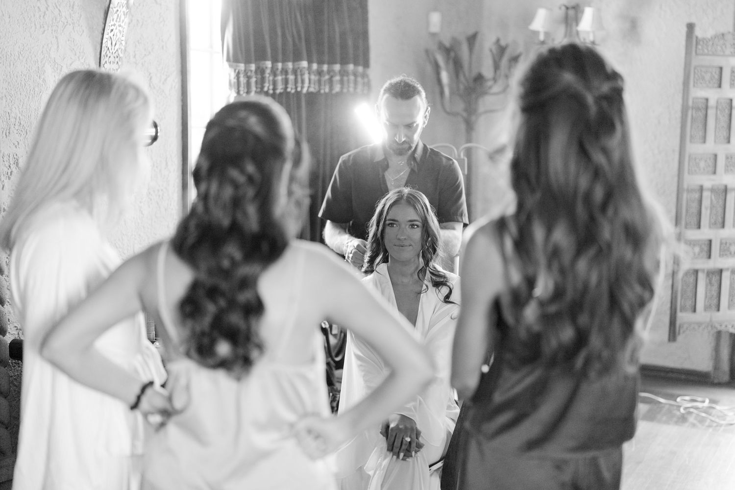 Black and white photo of wedding party gathering as bride prepares in ornate room with mirror.