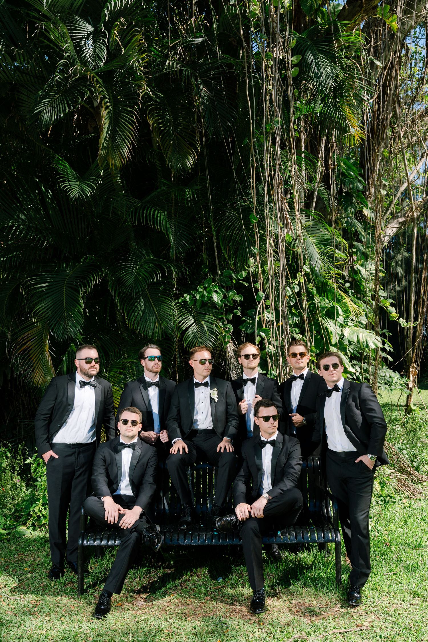 Eight groomsmen in black tuxedos and sunglasses pose outdoors on a bench surrounded by lush tropical greenery.