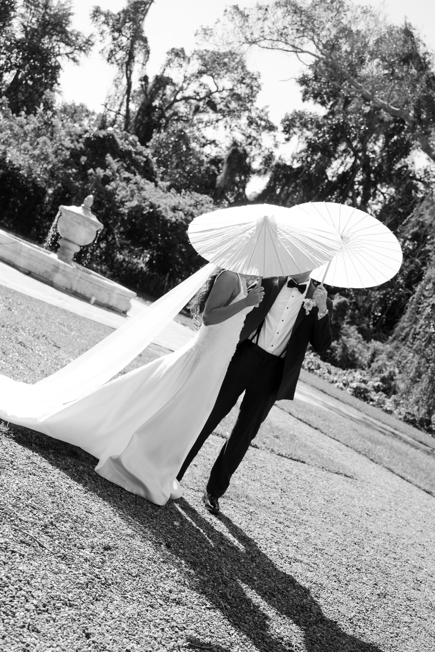 Black and white photo of bride and groom walking under white parasol with veil flowing behind.