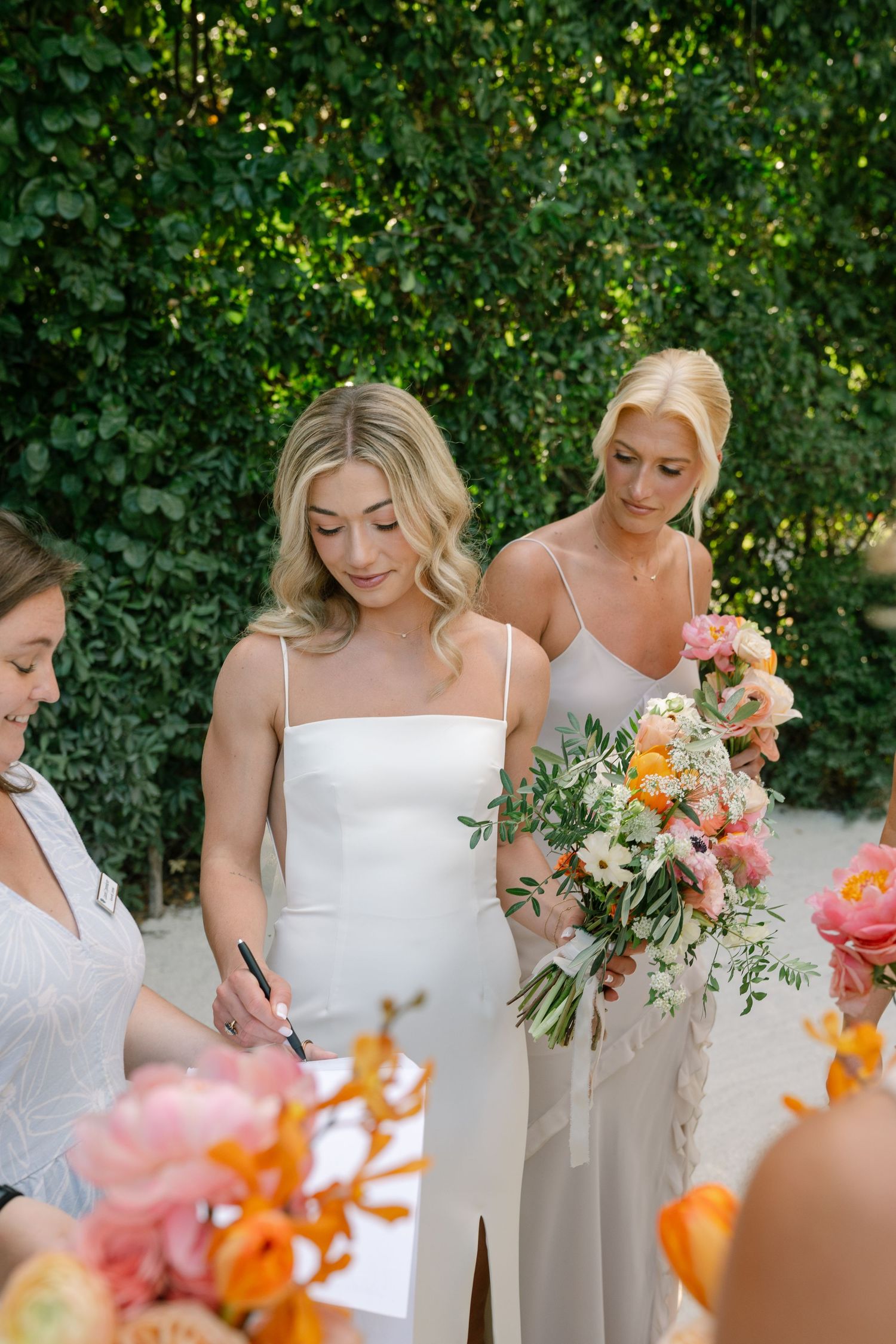 Bridesmaids in white dresses holding colorful tropical bouquets against green foliage.