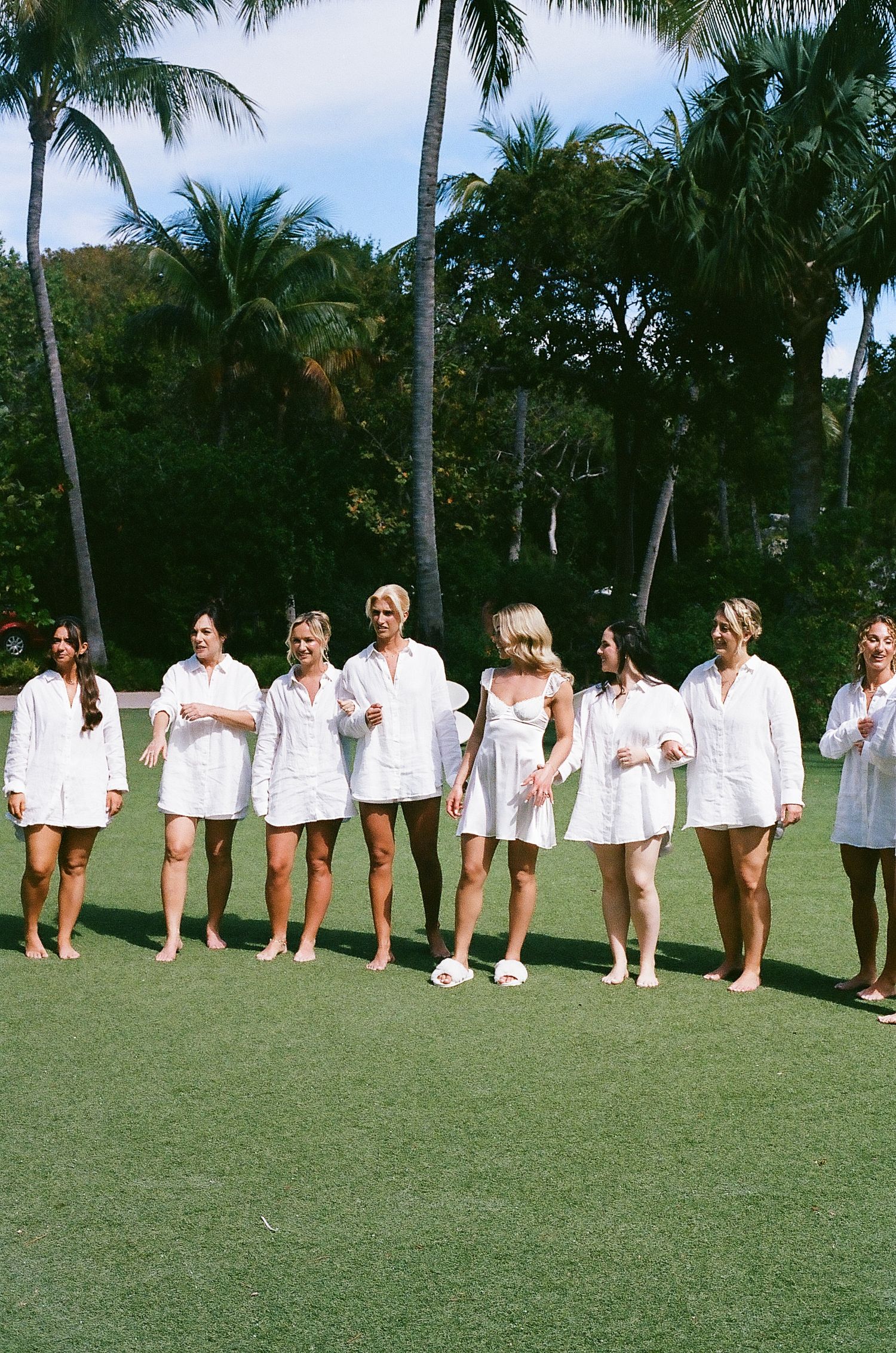 Bridesmaids in white dresses lined up on a tropical palm-lined lawn.