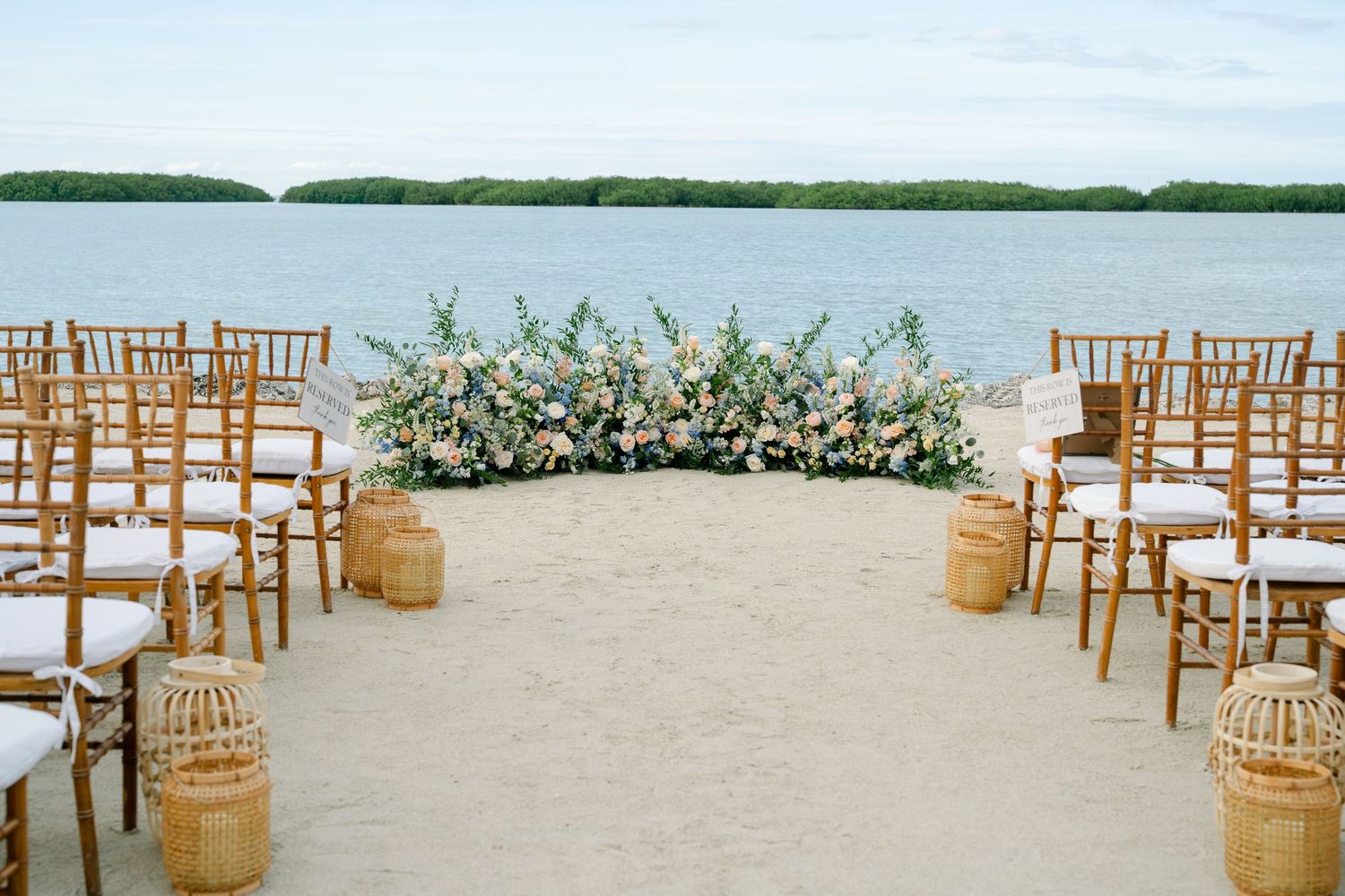 Elegant beach wedding ceremony setup with wooden chairs and floral arch overlooking calm waters.