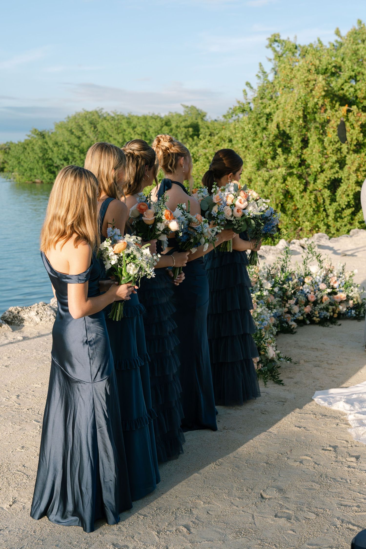 Bridesmaids in navy blue dresses holding floral bouquets pose on a tropical beach.