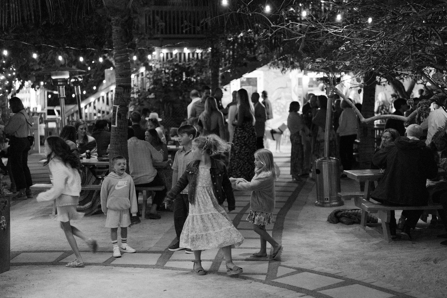 A lively outdoor evening dance gathering with couples moving to music under string lights in a park setting.