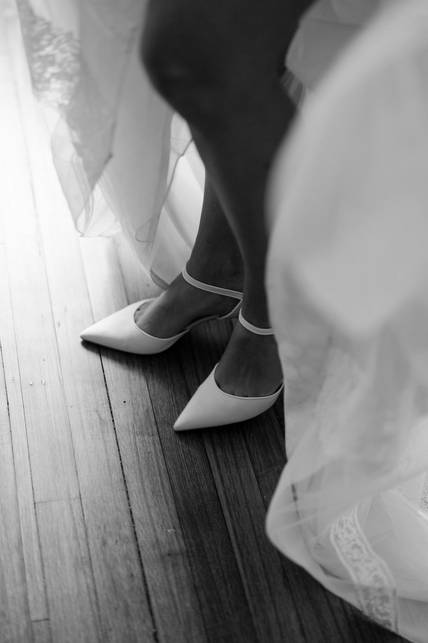 Elegant white bridal shoes peek out from beneath a flowing wedding dress on a dark hardwood floor.