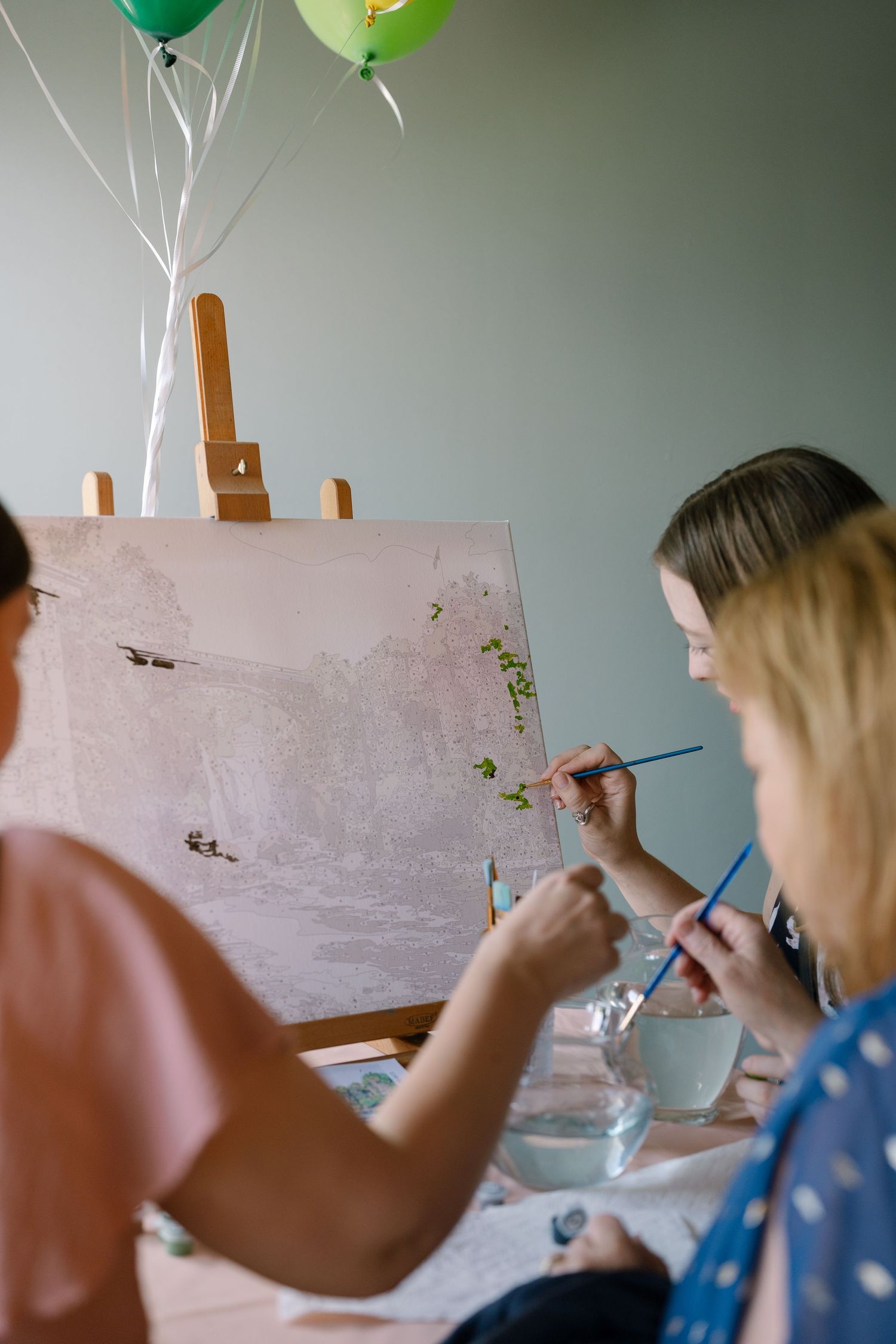 Artists paint together at a table with green balloons floating above and an easel displaying their canvas.