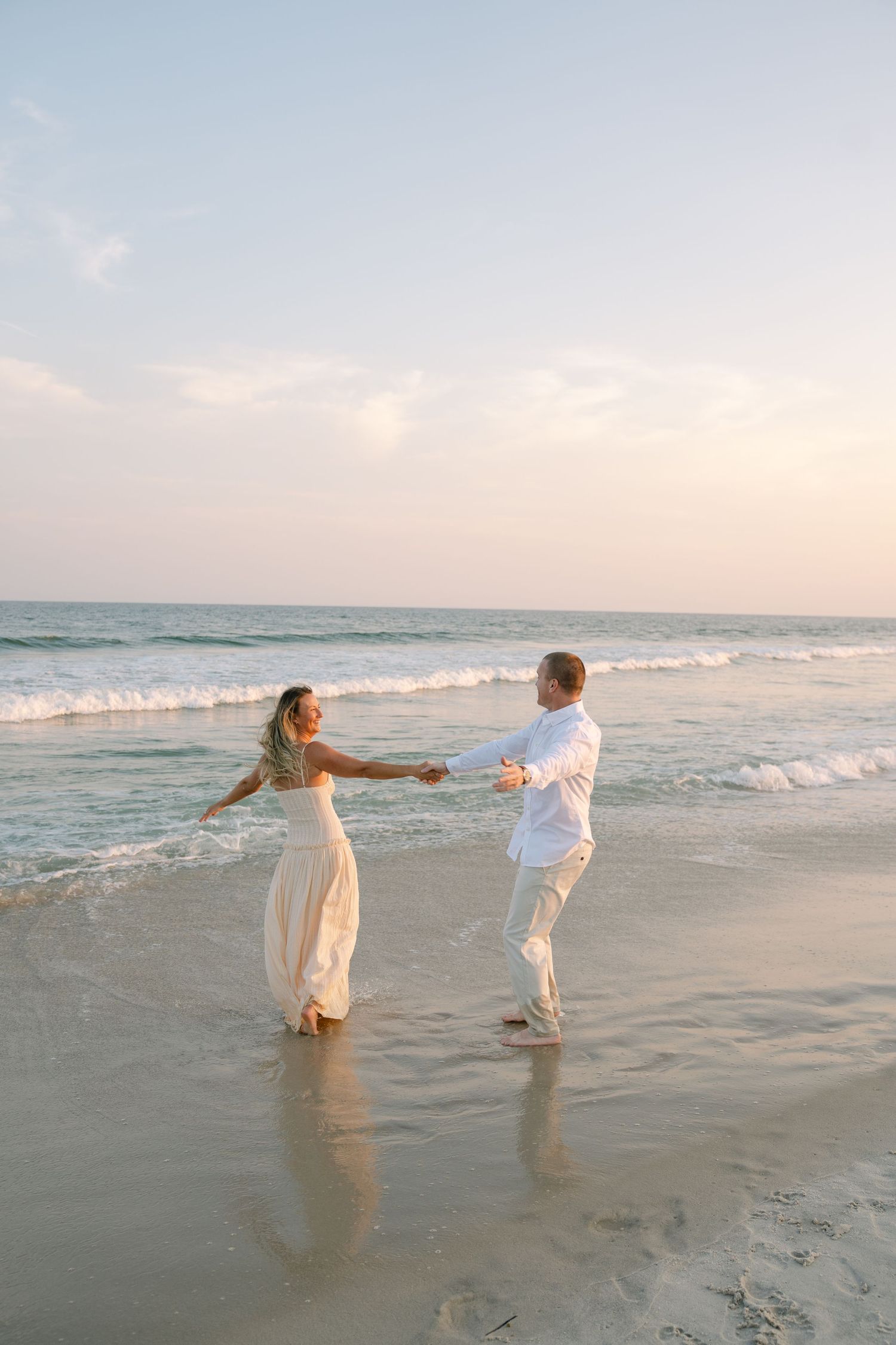 A couple in white outfits dances together on a beach at sunset with ocean waves rolling in the background.
