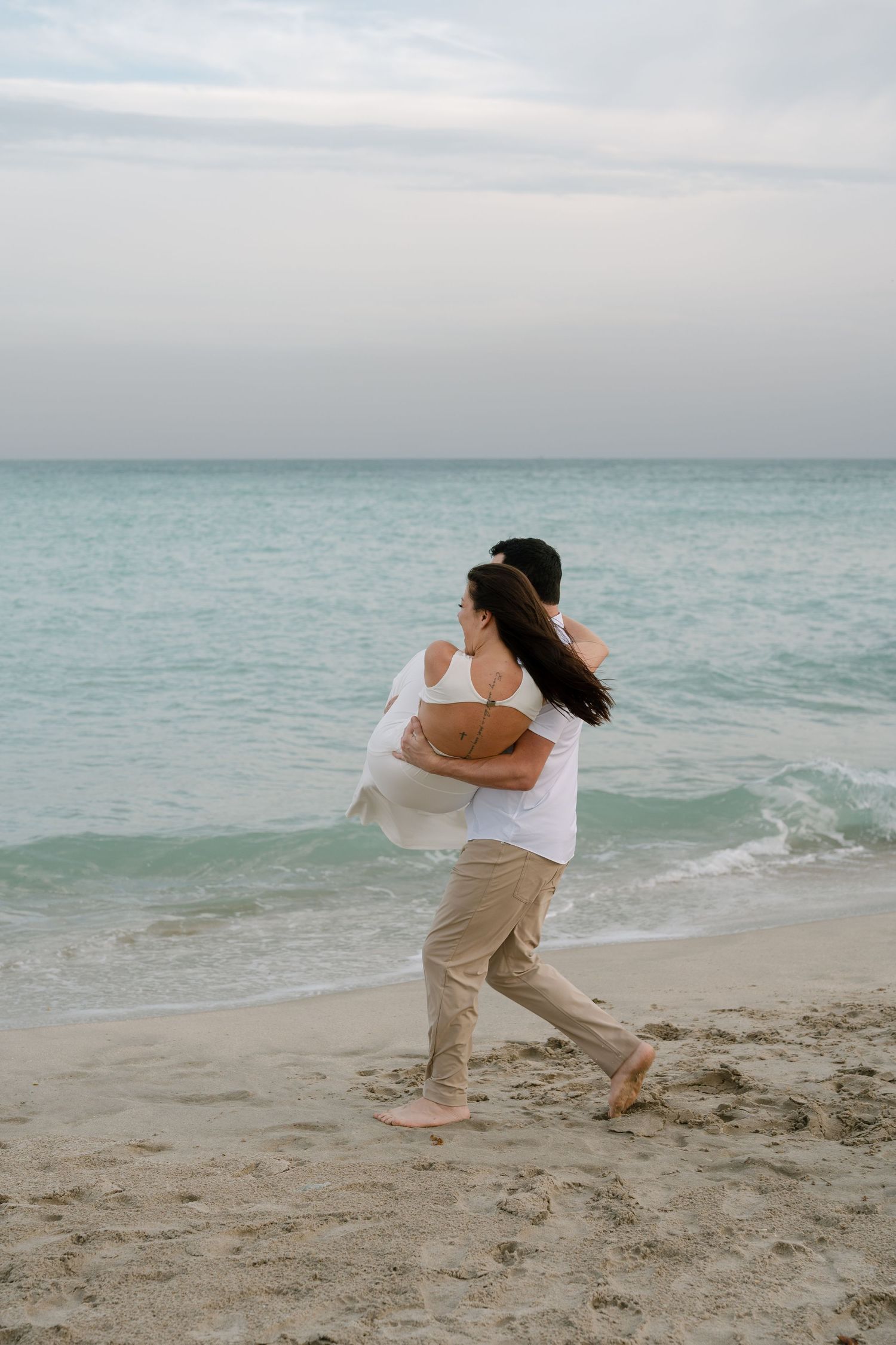 A couple dances playfully on a serene beach with gentle waves and overcast sky in the background.