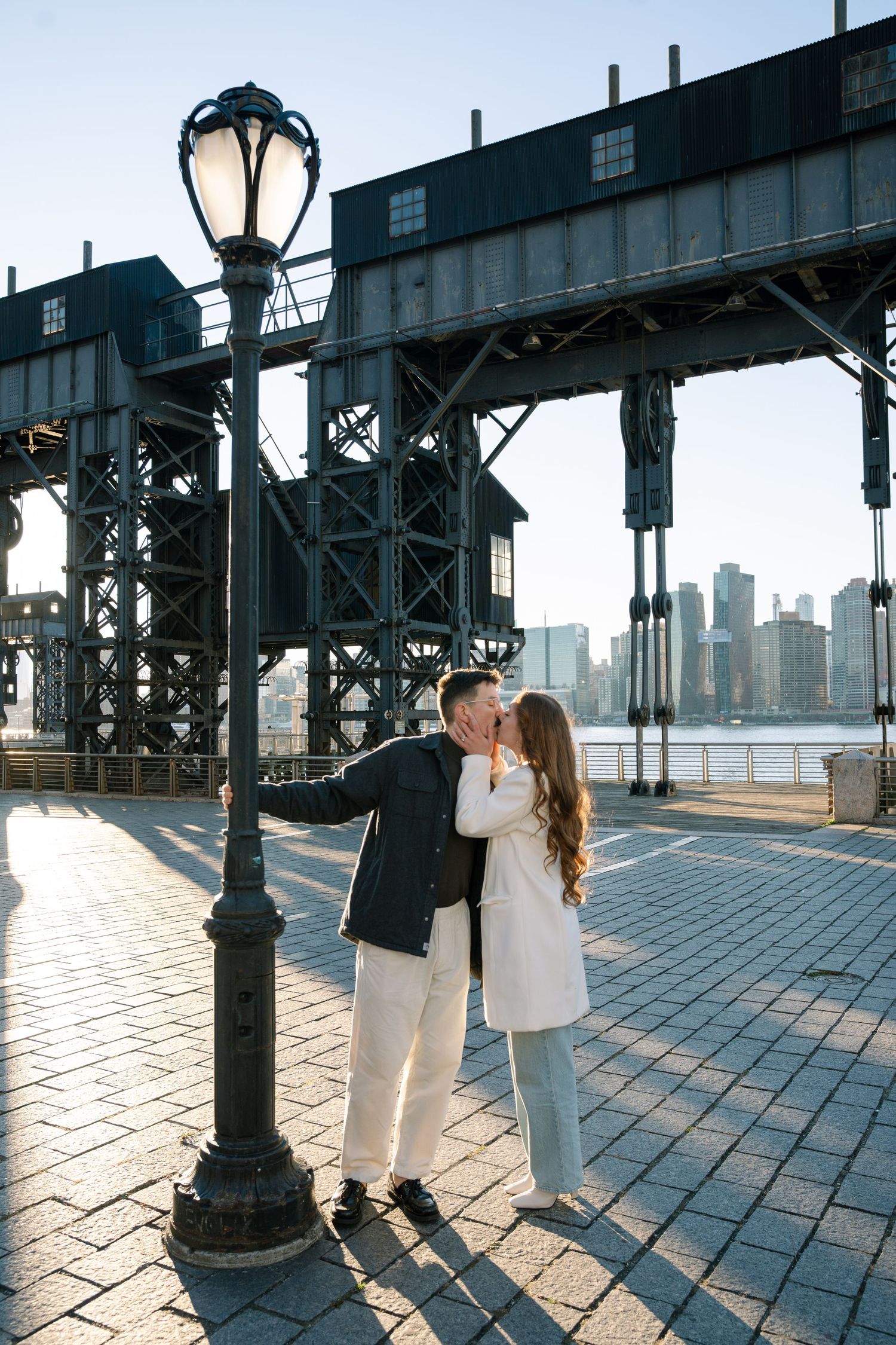 Romantic couple shares intimate moments along an urban boardwalk with city skyline views at sunset.