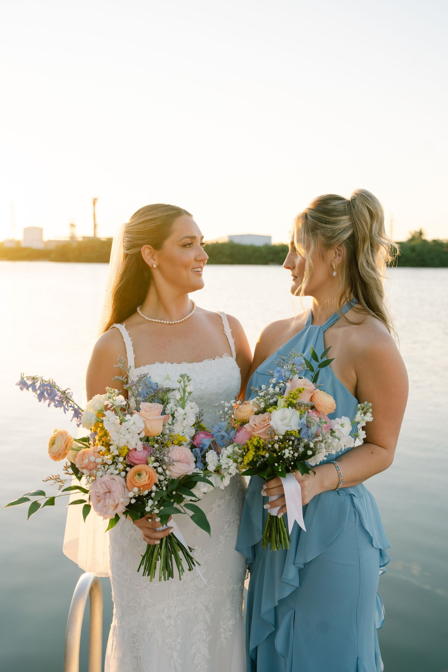 Two women in formal attire holding beautiful floral bouquets by the waterfront at sunset.