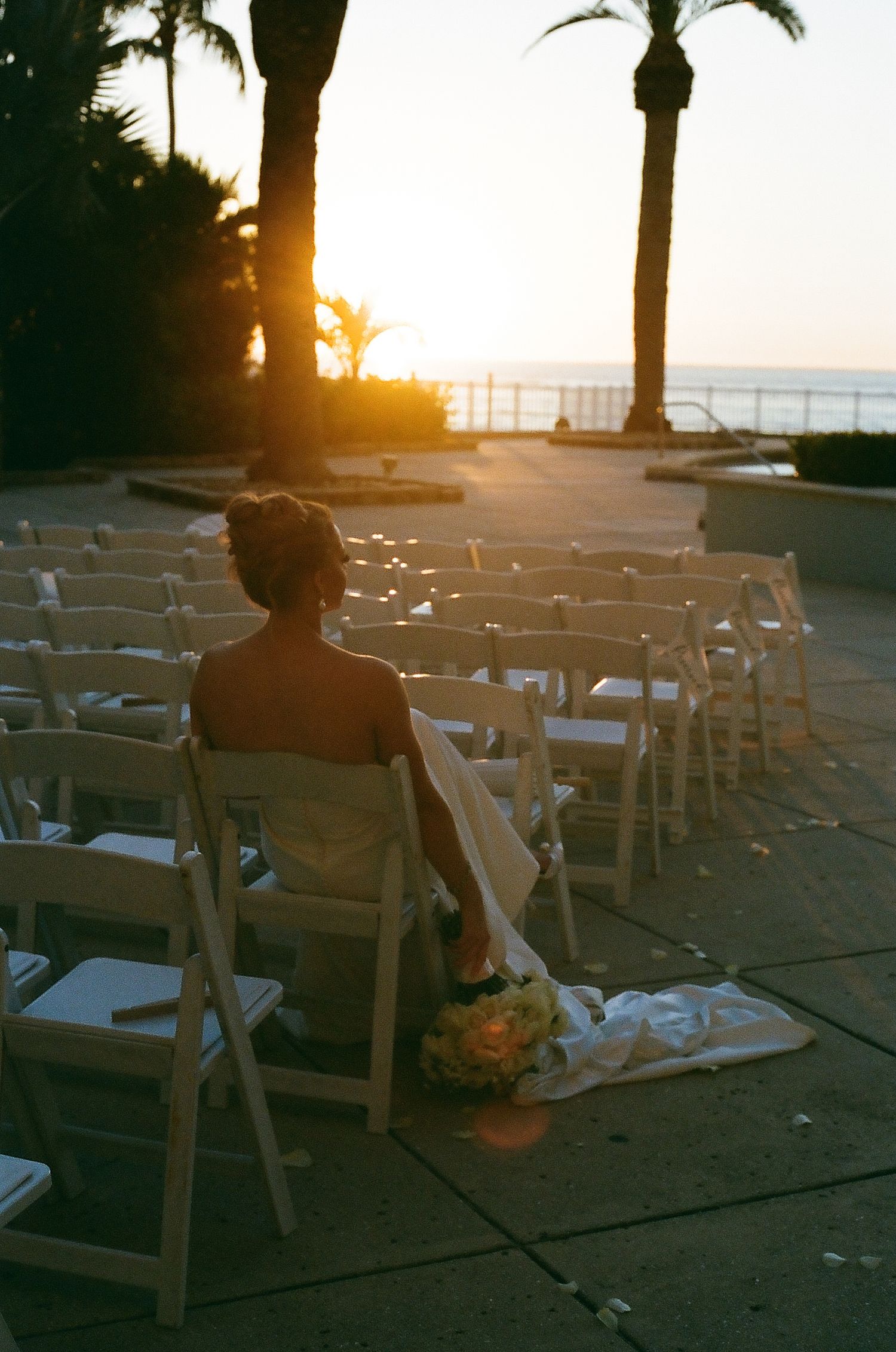 Empty white folding chairs set up for an outdoor sunset wedding ceremony with palm trees in the background.