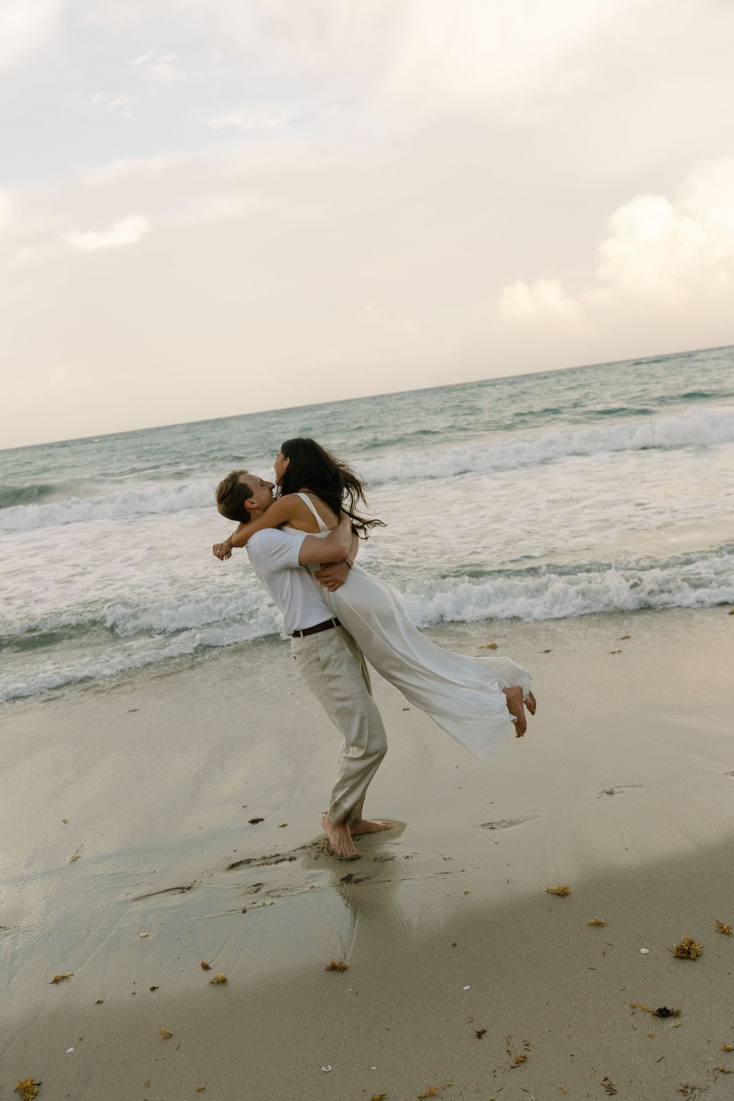 A couple shares a romantic embrace on a sandy beach, with ocean waves in the background, as the man lifts the woman in white.