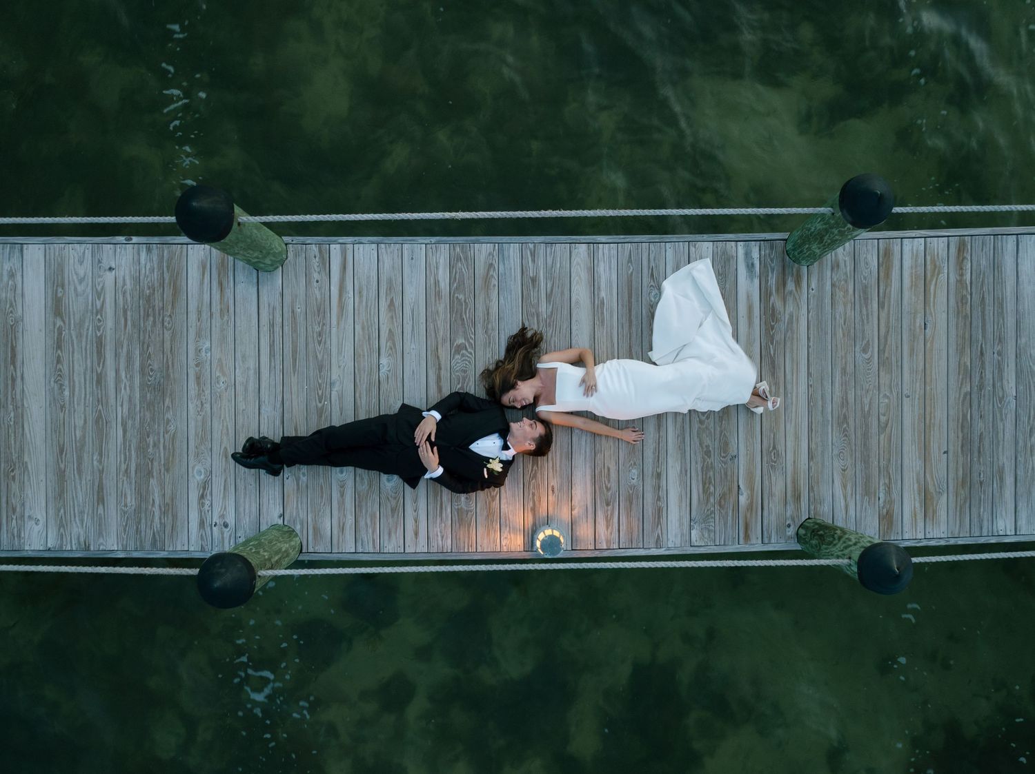 Aerial drone view of bride and groom lying on wooden dock surrounded by green water.