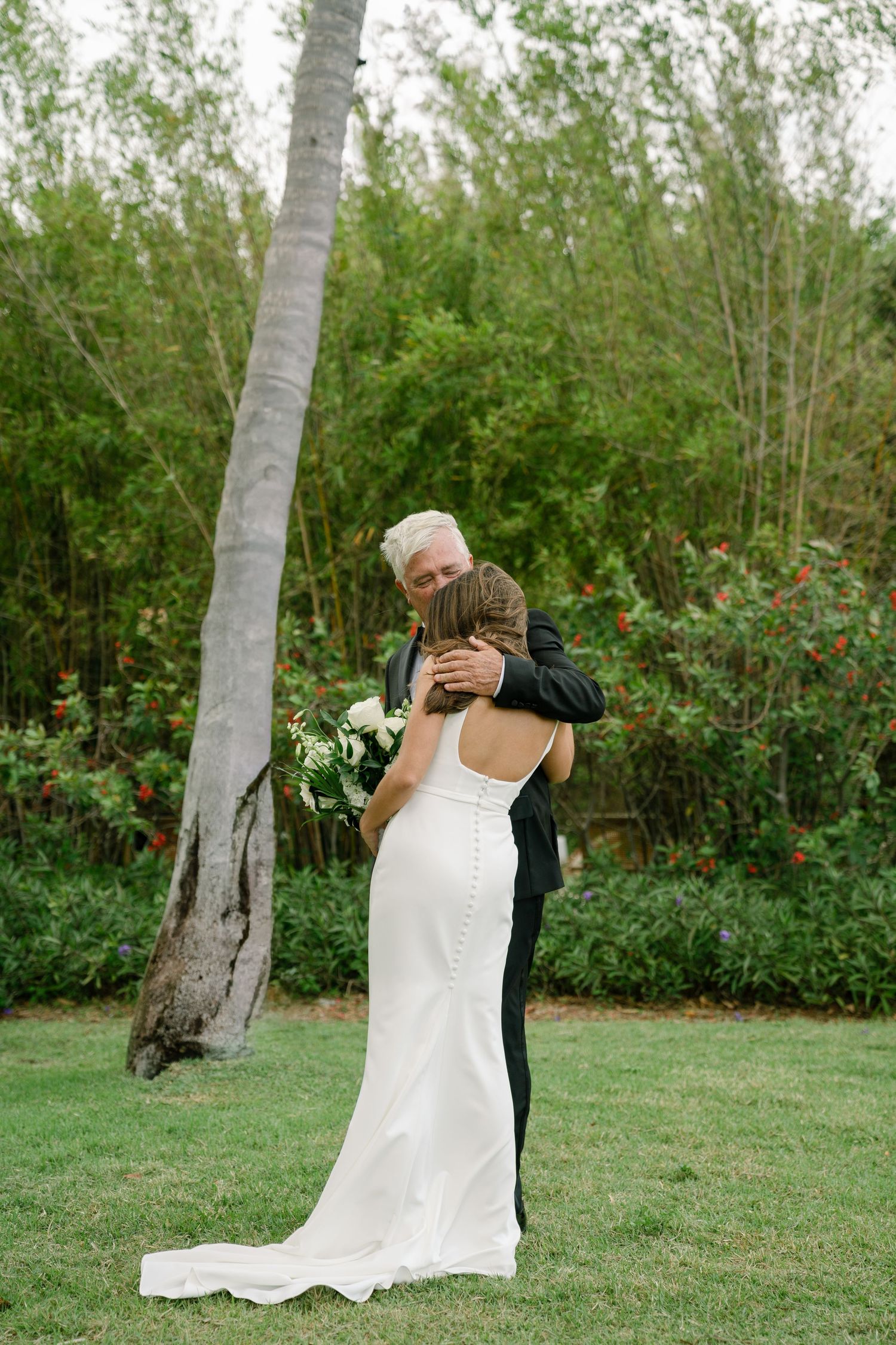 Bride in fitted white gown with button back embraces older gentleman in black suit outdoors near tropical palm tree.
