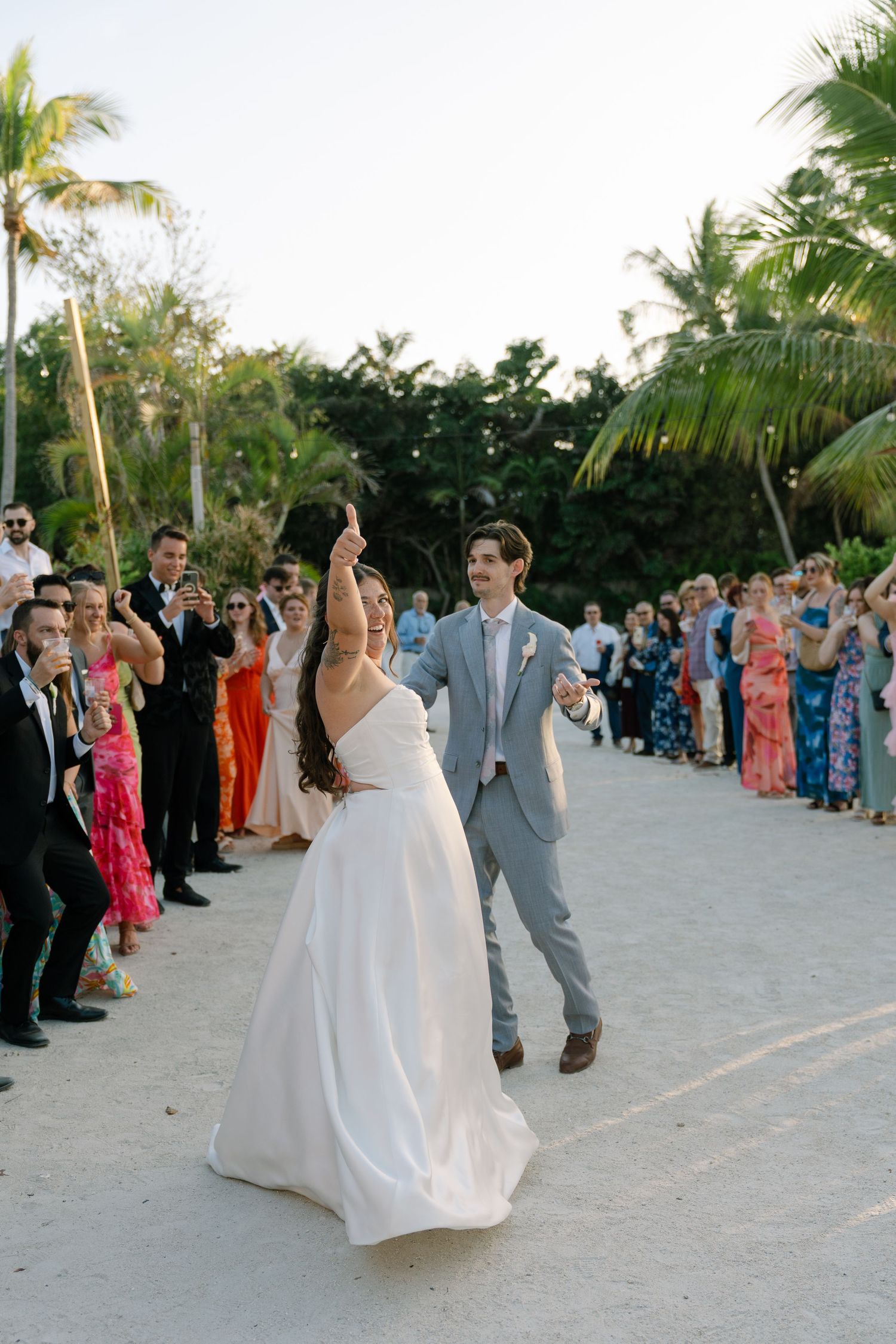 Bride and groom dancing on sandy beach at outdoor wedding reception surrounded by guests and palm trees.