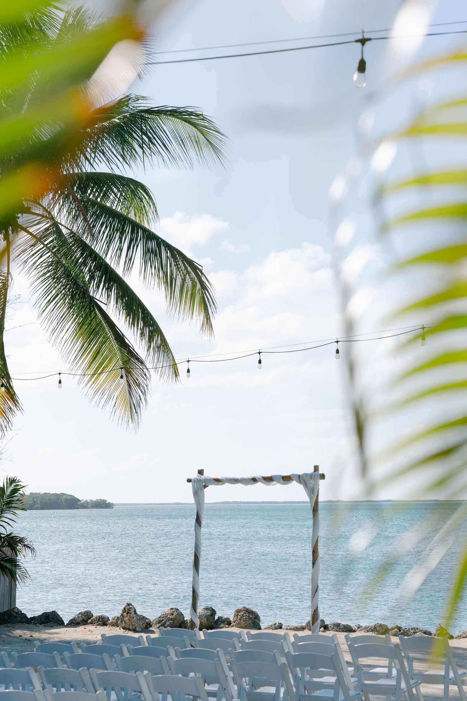 A tropical beach scene with palm trees, a wooden arch, and white chairs set up for an event or wedding.