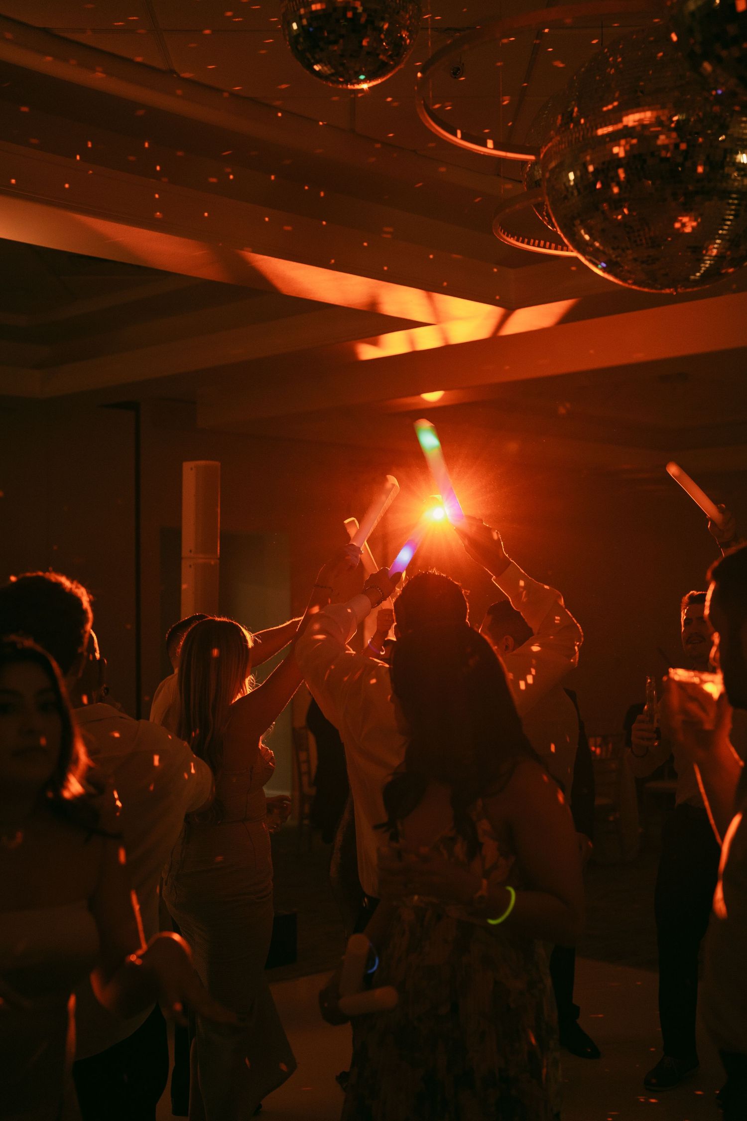 Silhouettes of people dancing under disco balls with warm orange lighting at a nightclub party.