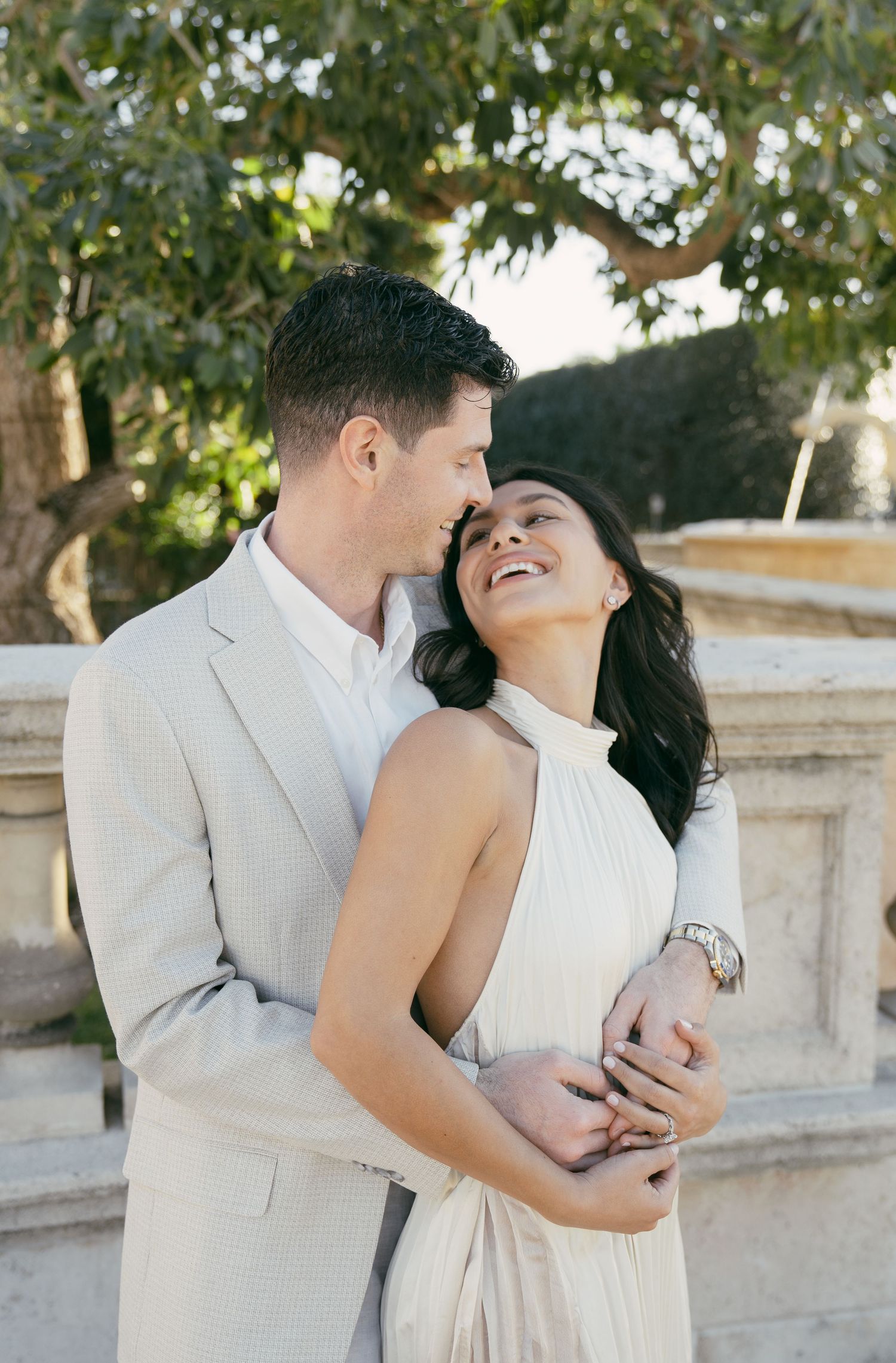 A couple in light-colored formal attire share a tender embracing moment against a stone wall backdrop in a sunlit garden setting.