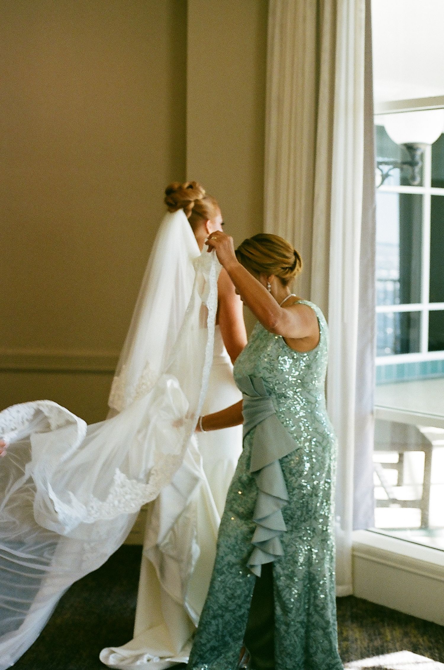 A figure in a mint green sequined dress helps adjust a flowing white wedding veil near a bright window.