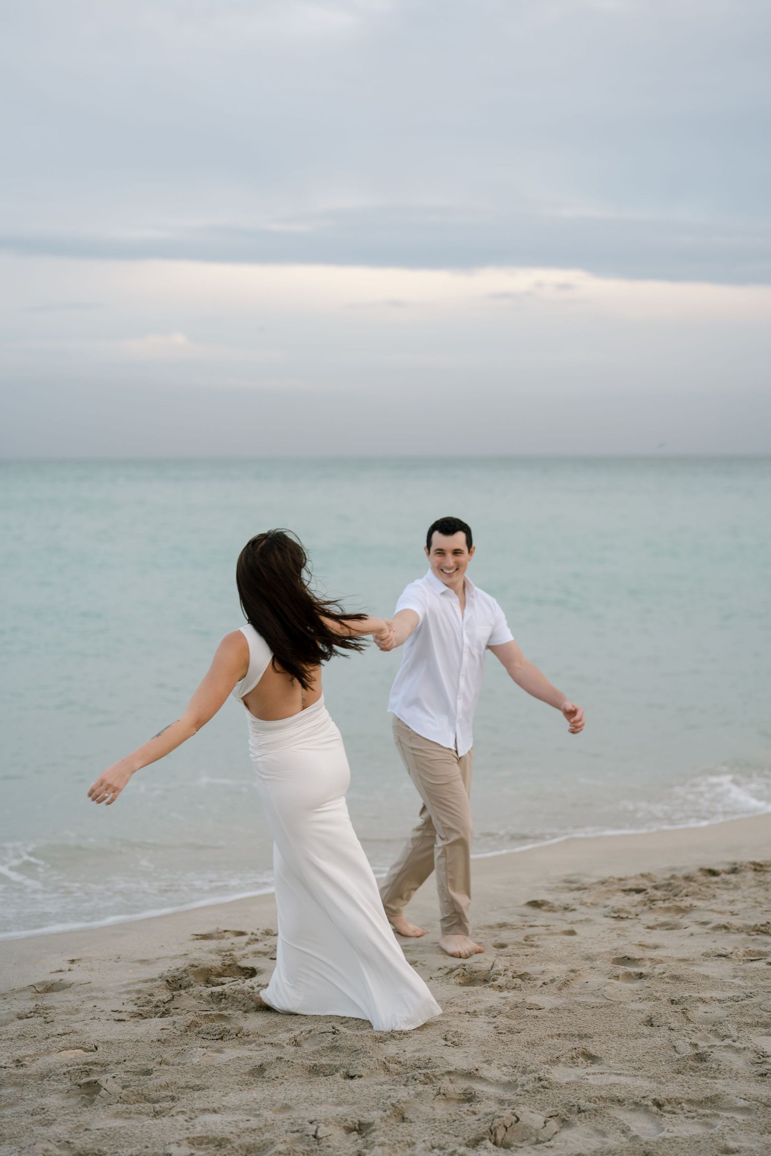 Couple in white and beige outfits dancing playfully along ocean shoreline at dusk.