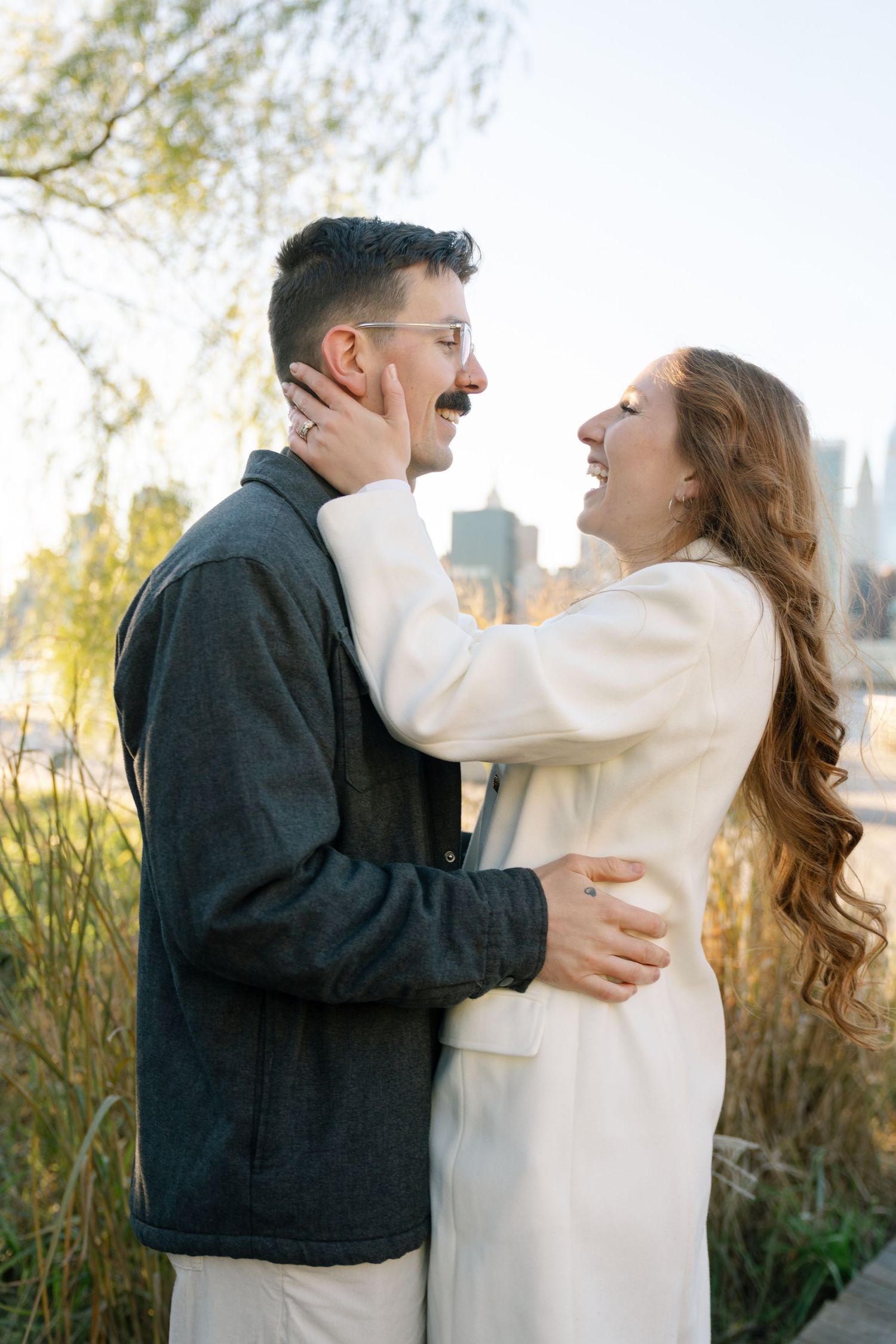 A couple embraces and laughs together outdoors during a romantic autumn photoshoot with trees in the background.