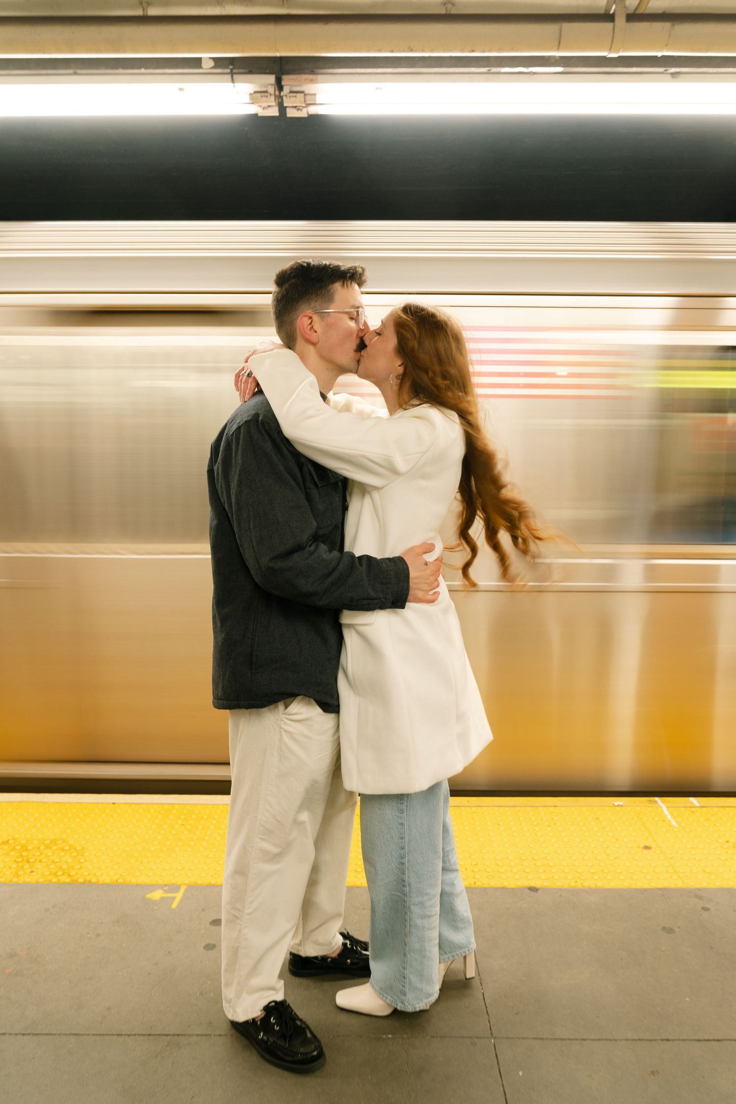 A couple embraces on a subway platform as a blurred train passes by in the background.