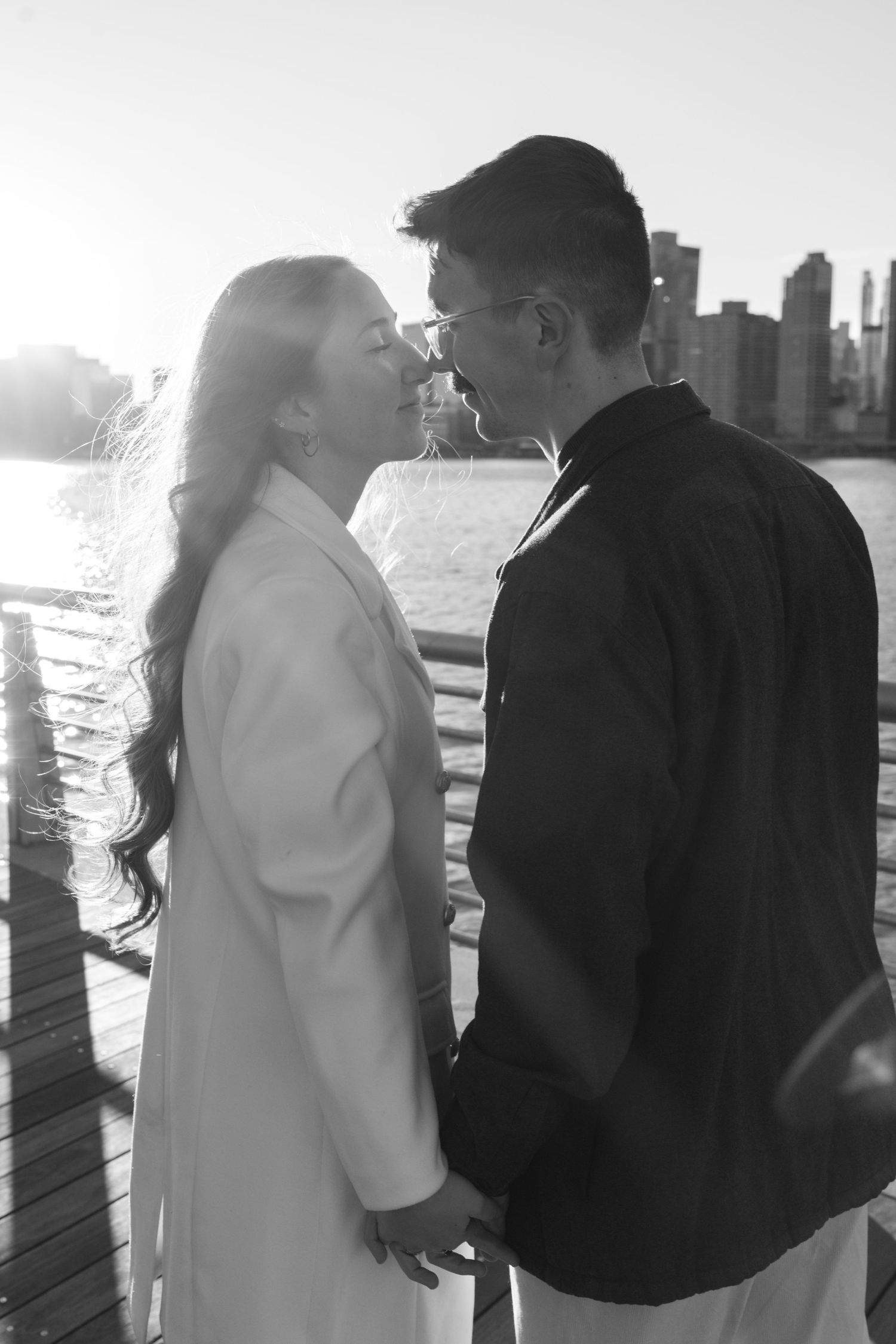 A romantic couple in black and white silhouette embraces on a pier with a cityscape in the background.