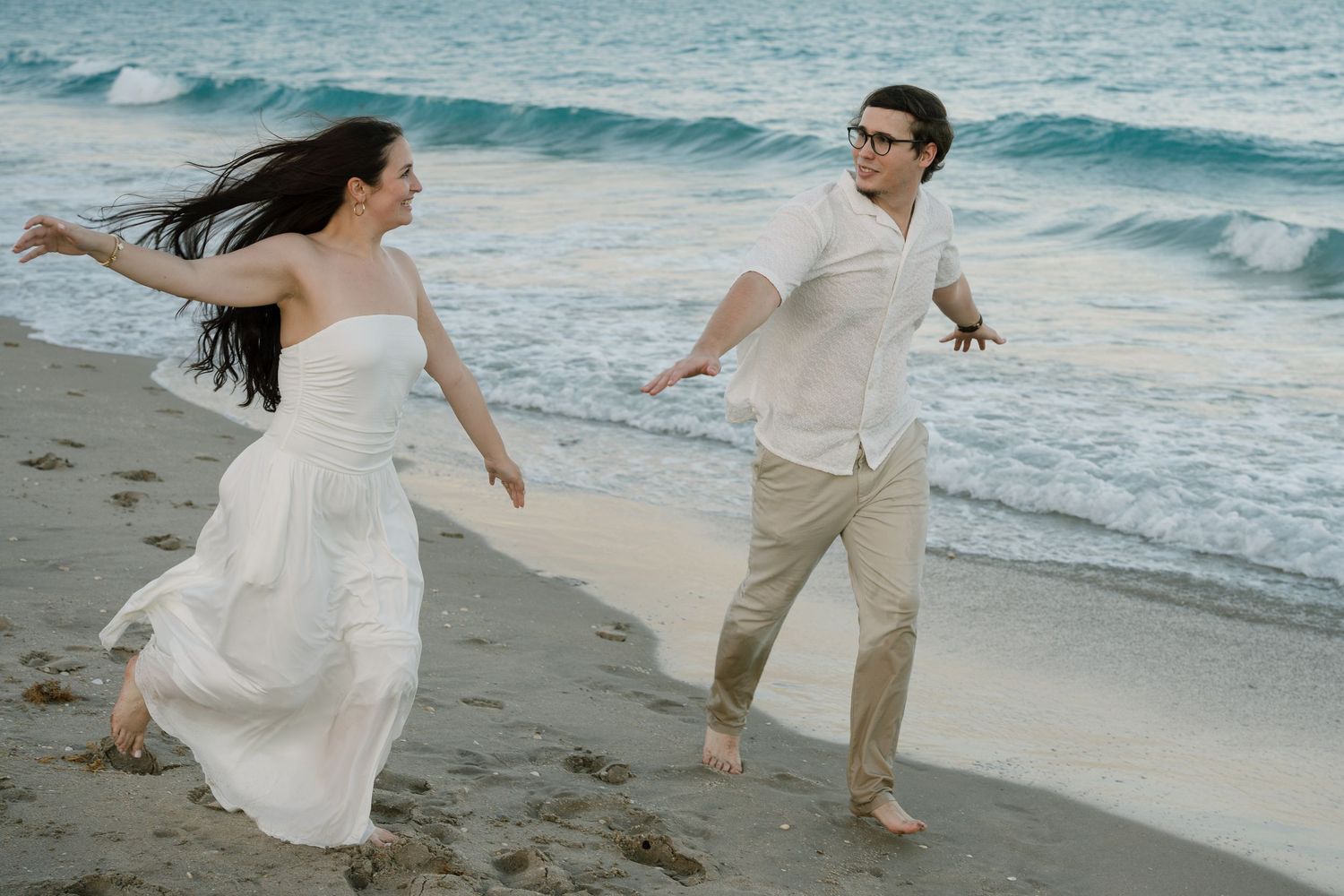 A couple playfully runs and dances together along a scenic beach with crashing waves and turquoise waters in the background.